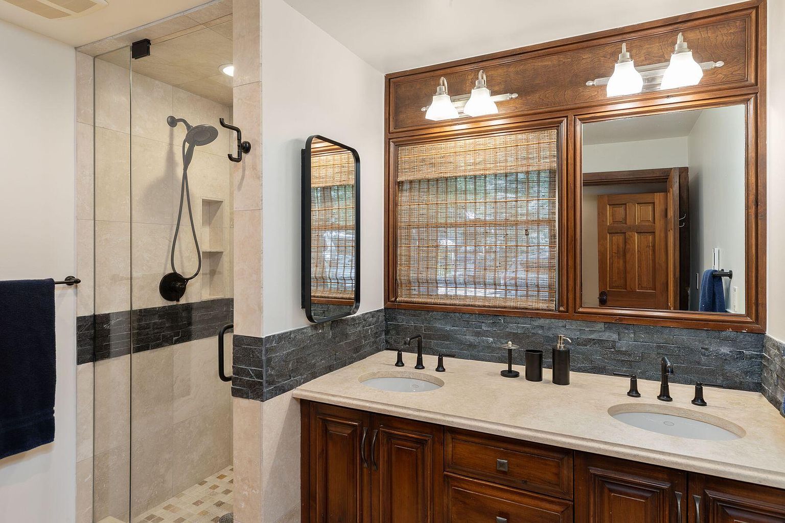 This is a well-appointed primary bathroom featuring a double vanity with dark wood cabinetry and a light-colored countertop. A large mirror with wood trim and decorative lighting hangs above the vanity, complemented by a smaller, rectangular mirror on the side. The shower is enclosed with glass and features a dark showerhead and tiled walls, adding a touch of modern elegance.