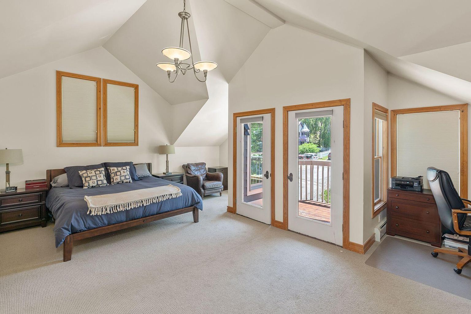 This is a primary bedroom featuring a king-sized bed with blue bedding and decorative pillows. The room has a neutral color palette with white walls and light carpet. Natural light floods the space through two windows and two sets of French doors leading to a balcony, creating a bright and airy atmosphere. A desk area is visible to the right, and a leather chair is in the corner.