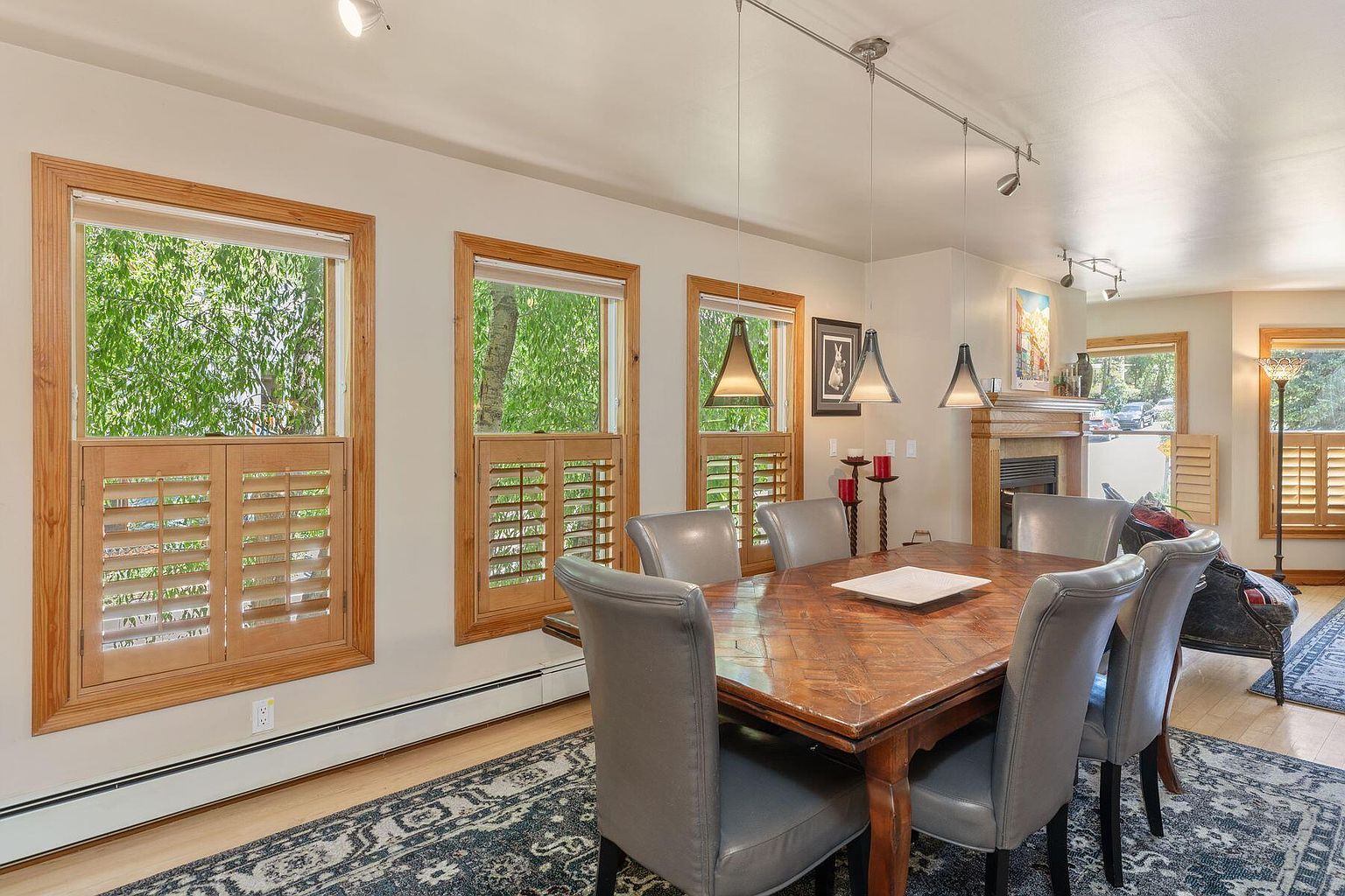 This is a dining room featuring a wooden table with gray upholstered chairs, set on a patterned rug. Three windows with wooden shutters provide natural light, and pendant lights hang above the table. A fireplace is visible in the background, adding a cozy touch to the room.