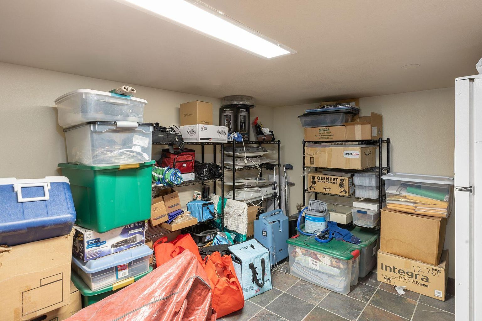 The image showcases a cluttered basement storage area. Shelving units are packed with boxes, bins, and various household items. The floor is tiled, and a fluorescent light fixture illuminates the space, giving the impression of a functional but disorganized storage solution.