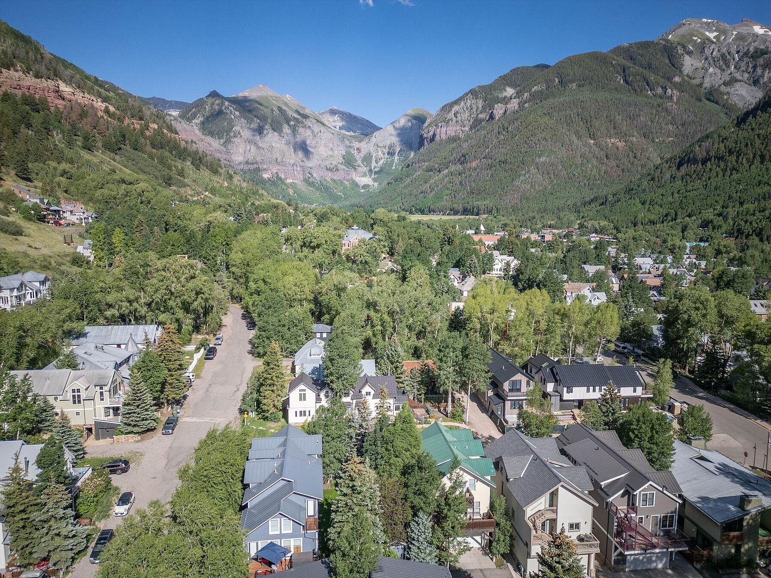 This aerial shot showcases a charming town nestled in a valley surrounded by majestic mountains. The houses feature a mix of architectural styles, with well-maintained yards and mature trees providing ample shade. The overall impression is one of a peaceful and picturesque community.