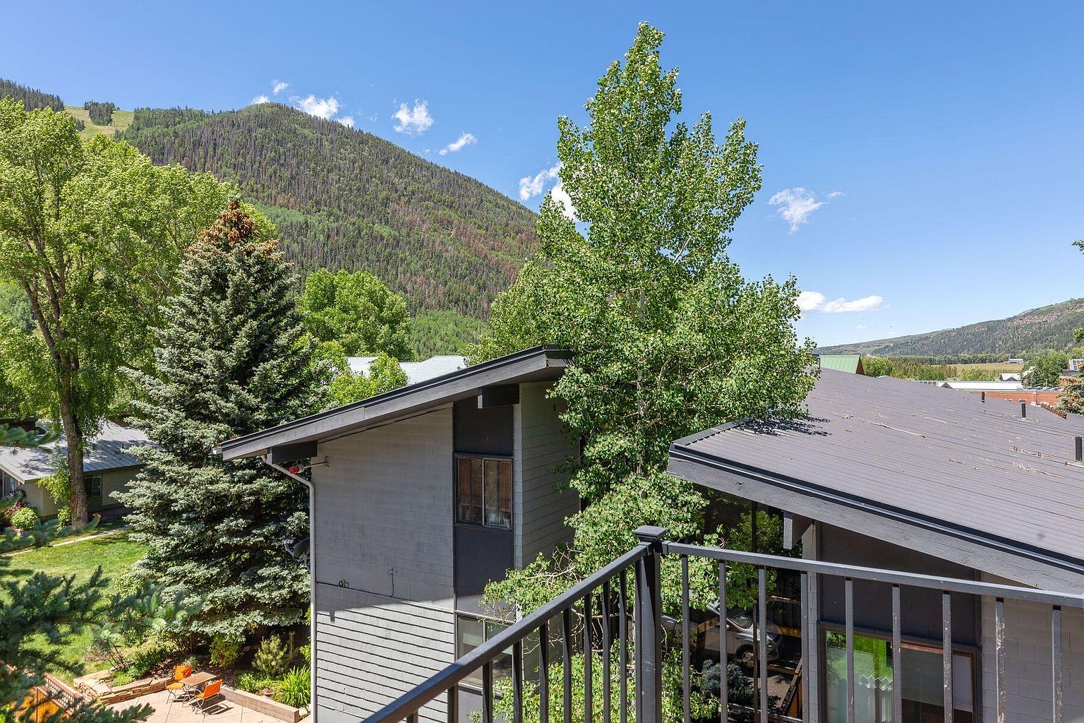 This image showcases a balcony view with a mountain backdrop. The balcony features a black metal railing, and the architecture includes modern, gray-toned buildings with dark roofs. Lush greenery and trees surround the buildings, creating a serene and natural setting.