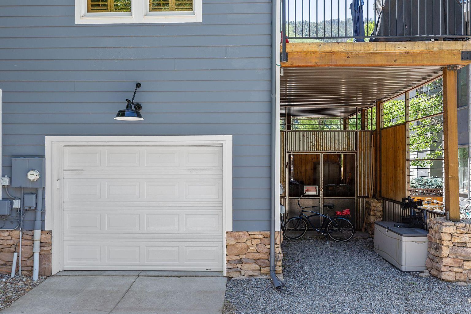 The image showcases the exterior of a garage with a white paneled door, complemented by a blue-gray siding. Stone accents are visible at the base of the structure, adding a touch of rustic charm. To the right, there's a covered area with a bicycle and storage container, suggesting functional outdoor space.