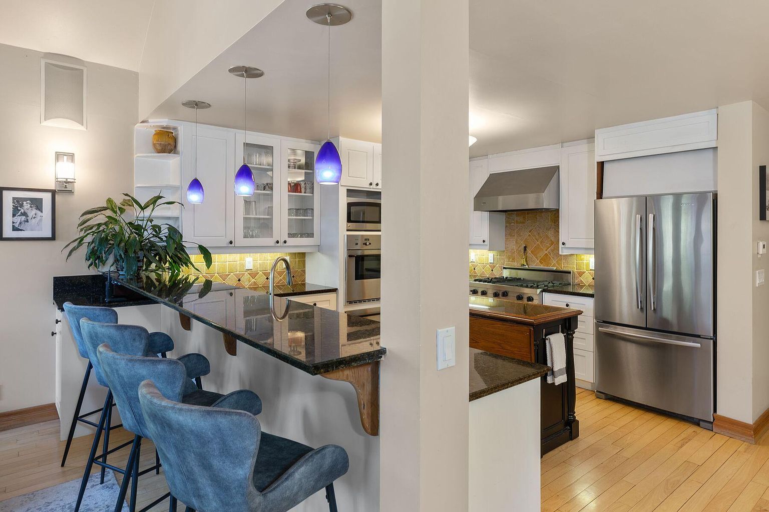 This is a well-lit kitchen featuring white cabinetry, stainless steel appliances, and a dark countertop breakfast bar with seating. The kitchen includes a stainless steel refrigerator, oven, and range hood. The flooring is hardwood, and the overall style is modern and functional.