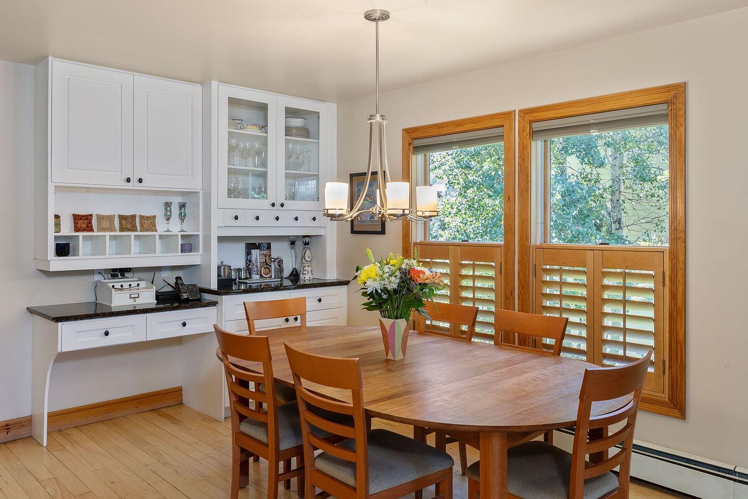 This is an inviting dining room featuring a large, oval wooden table surrounded by six wooden chairs with gray cushions. Natural light floods the room through two windows with wooden shutters, complementing the warm tones of the hardwood flooring. A built-in hutch with white cabinetry and a dark countertop adds functionality and style to the space.