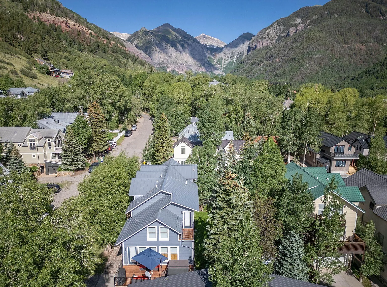 This aerial shot captures a picturesque neighborhood nestled amidst lush greenery and majestic mountains. The houses feature varying architectural styles, with visible rooftops and well-maintained exteriors. It gives a sense of the community's peaceful setting and the natural beauty surrounding the properties.