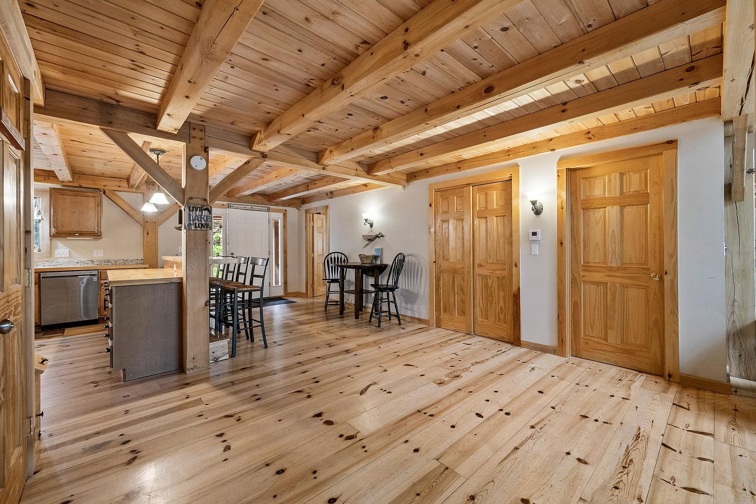 This open-concept interior features a rustic aesthetic with exposed timber beams on the ceiling and light wood flooring throughout. The space includes a functional kitchen area with a breakfast bar, a small dining nook with a table and chairs, and multiple wooden doors leading to other rooms. The warm, natural wood tones and open layout create a cozy, inviting atmosphere typical of a cabin or lodge-style home.
