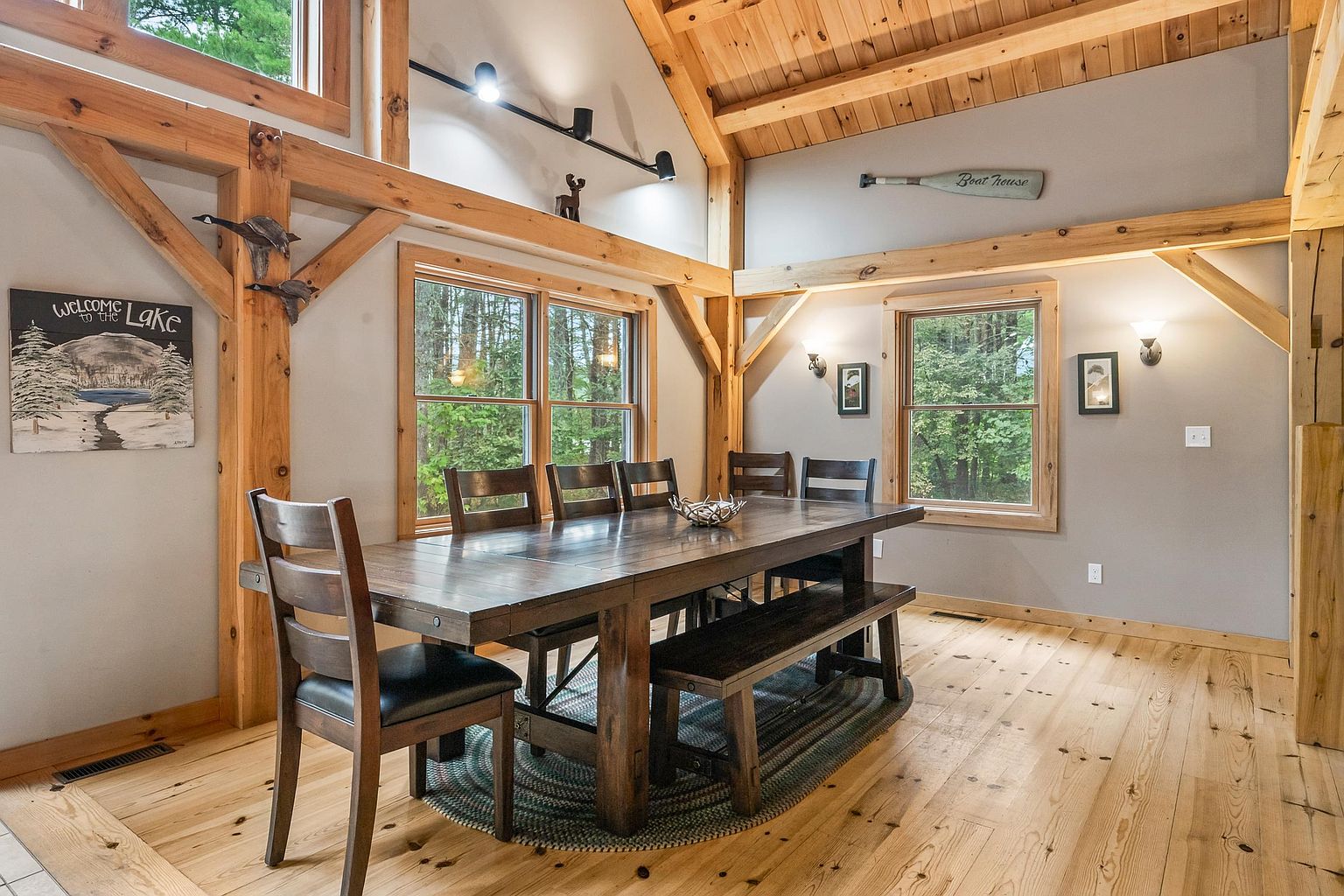This rustic dining room features a large wooden table with matching chairs and a bench, set upon a braided oval rug. The space is defined by exposed timber beams, light wood flooring, and neutral-toned walls, creating a warm, cabin-like atmosphere. Large windows allow natural light to fill the room, which is accented by decorative wall art and a boat oar mounted above the window.