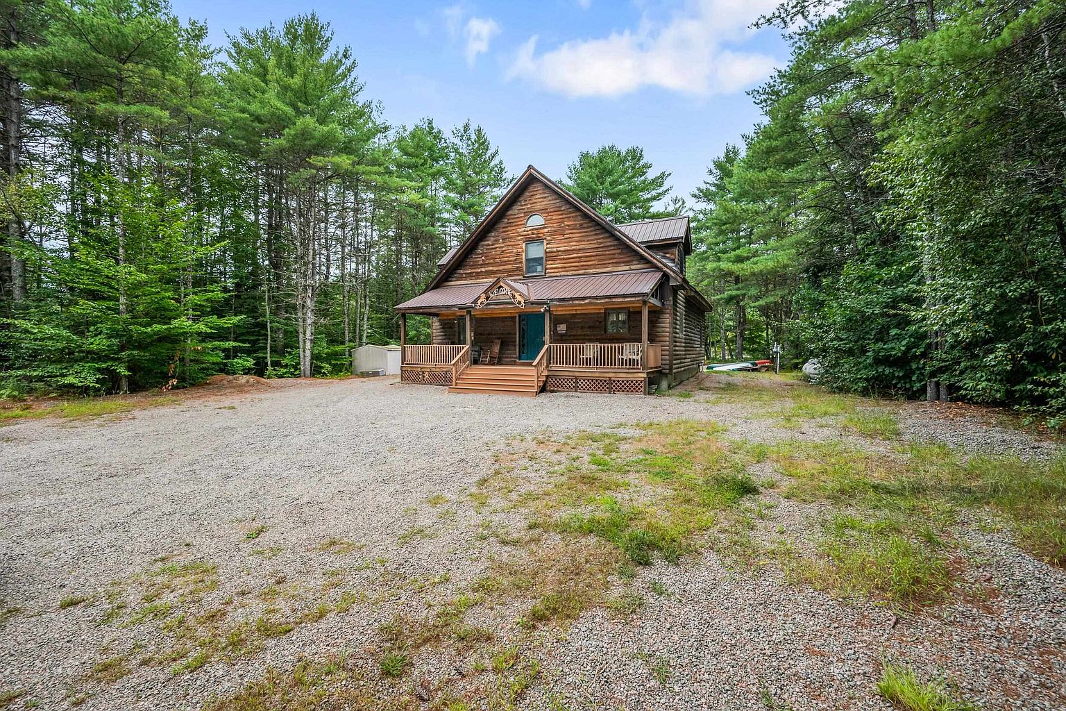 This charming log cabin features a prominent front porch with wooden railings and steps, set against a backdrop of dense, mature pine trees. The exterior is constructed from horizontal logs, complemented by a dark metal roof and a central window on the upper level. A wide, gravel driveway leads directly to the home, creating a secluded and rustic atmosphere typical of a woodland retreat.