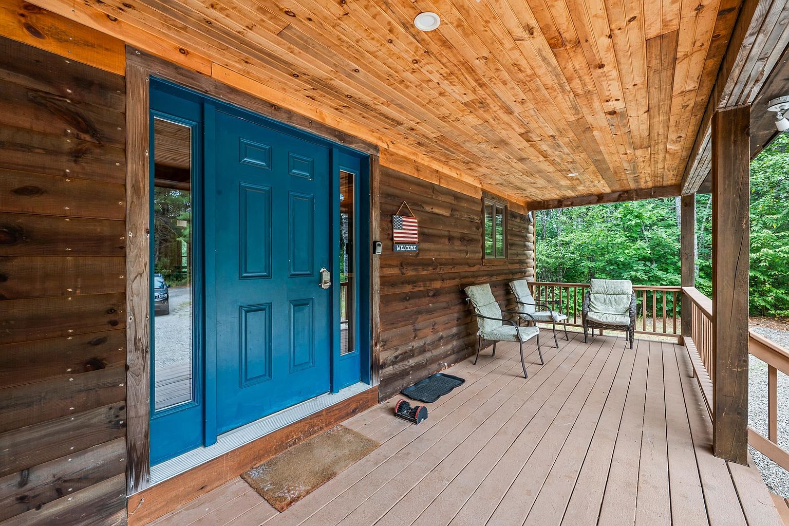 This inviting front porch features rustic log-style siding and a warm, natural wood-paneled ceiling. A vibrant blue front door with flanking sidelights serves as the focal point, complemented by a welcoming American flag sign and comfortable outdoor seating. The perspective captures the length of the covered deck, highlighting the peaceful, wooded surroundings visible through the wooden railing.