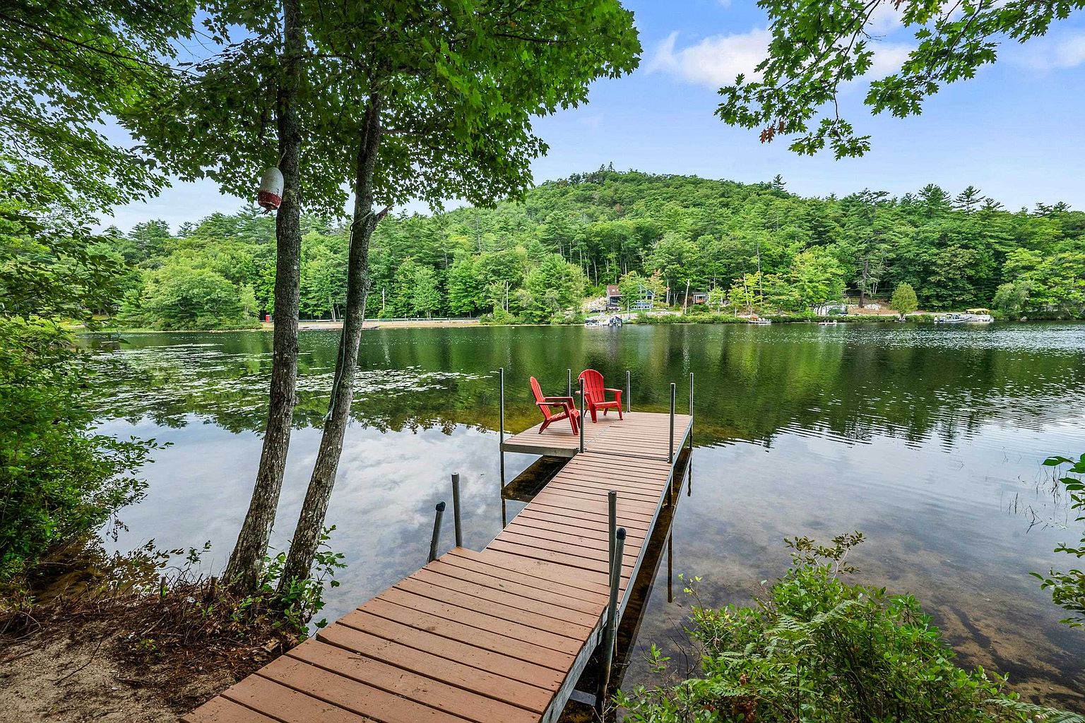 This serene waterfront scene features a wooden dock extending into a calm, reflective lake, framed by lush green trees and a gentle hillside in the background. Two bright red Adirondack chairs sit at the end of the dock, creating a peaceful spot for relaxation. The perspective is from the shoreline, looking out over the water, capturing the natural beauty and tranquil atmosphere of the property's outdoor space.