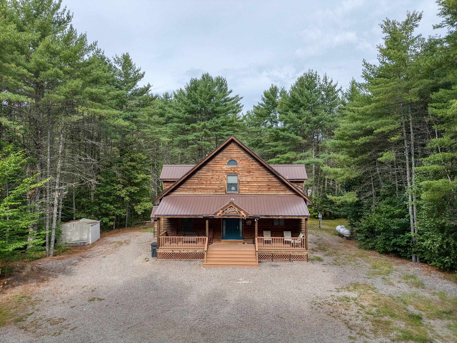 This image presents a front-facing view of a rustic log cabin nestled within a dense forest of tall pine trees. The home features a prominent A-frame roofline, a covered front porch with wooden railings, and a gravel driveway leading up to the entrance. The setting offers a secluded, tranquil atmosphere, emphasizing the natural beauty and woodland surroundings of the property.