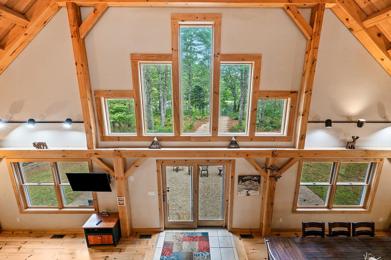This high-angle shot captures a spacious, rustic-style living area featuring exposed timber framing and a dramatic, multi-paneled window arrangement that frames the wooded exterior. The room includes a central glass-paneled door leading to the outdoors, flanked by large windows on either side, and is accented by decorative wildlife-themed decor on the timber beams. The warm wood tones and open-concept design create an inviting, lodge-like atmosphere perfect for a mountain or lake retreat.