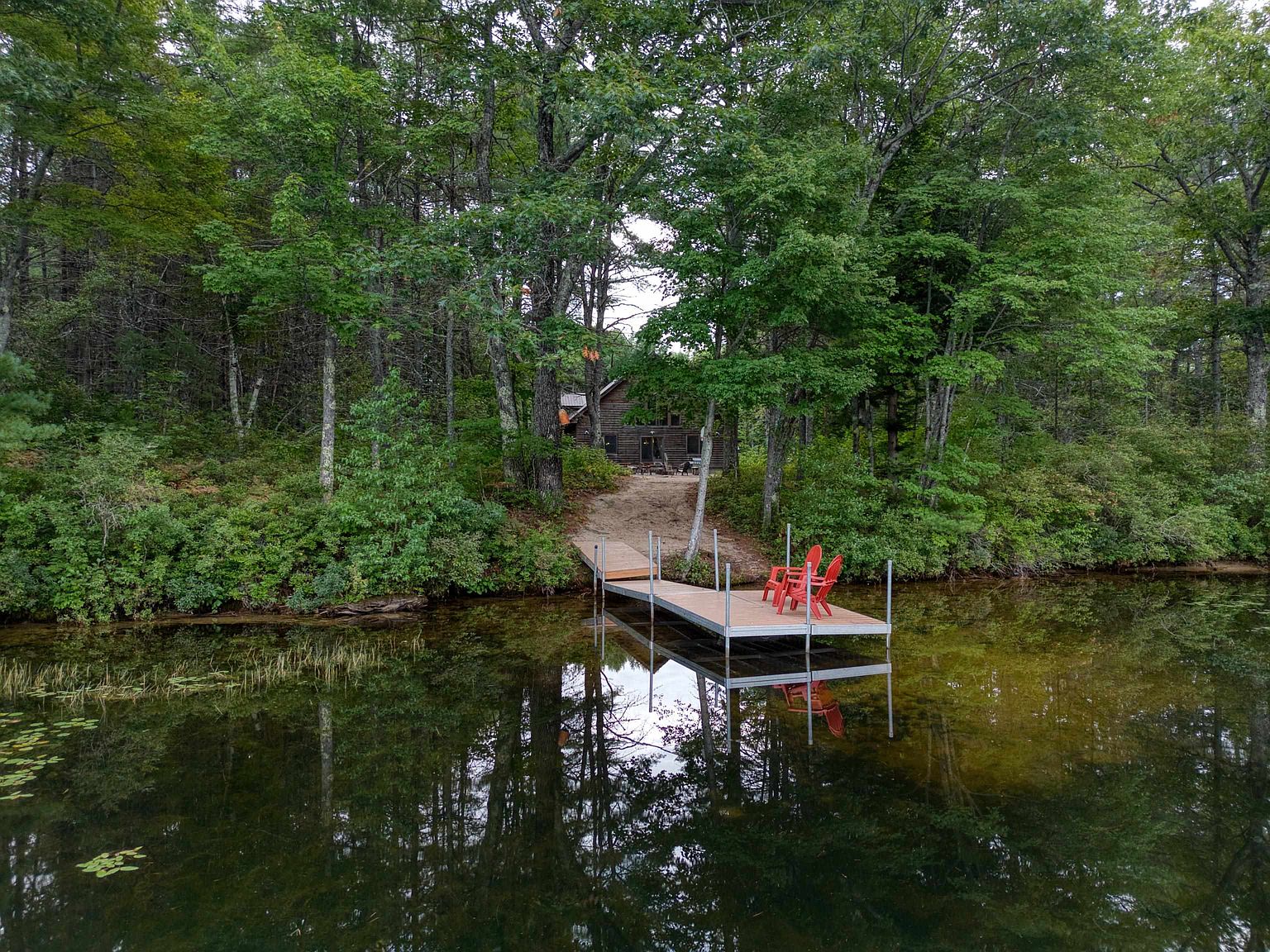 This aerial perspective captures a secluded lakeside cabin nestled within a dense, lush forest. A wooden dock extends from the shoreline into the calm, reflective water, featuring two bright red Adirondack chairs that provide a vibrant focal point. The scene evokes a peaceful, private retreat atmosphere, highlighting the natural beauty and direct water access of the property.