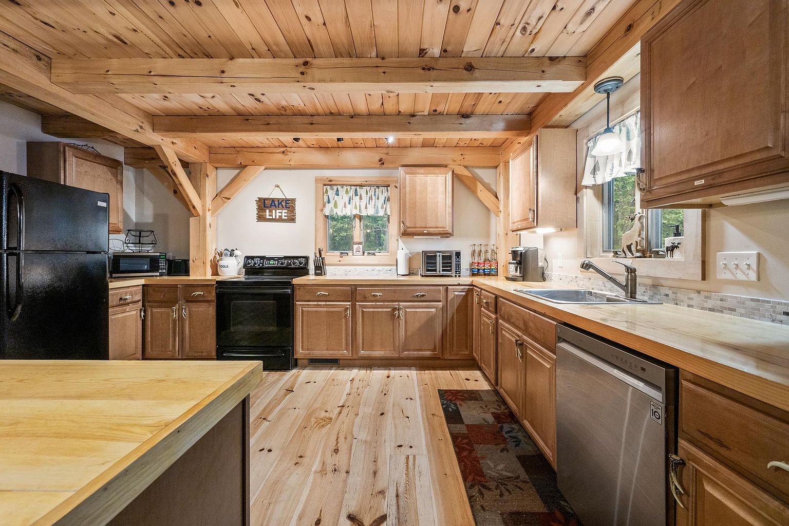 This rustic-style kitchen features warm wood cabinetry, light-toned wood flooring, and exposed timber ceiling beams that create a cozy, cabin-like atmosphere. The space is equipped with black appliances, a stainless steel dishwasher, and light-colored countertops, with a window over the sink providing natural light. A central island with a light wood surface anchors the room, while a decorative 'Lake Life' sign adds a charming, thematic touch to the wall.