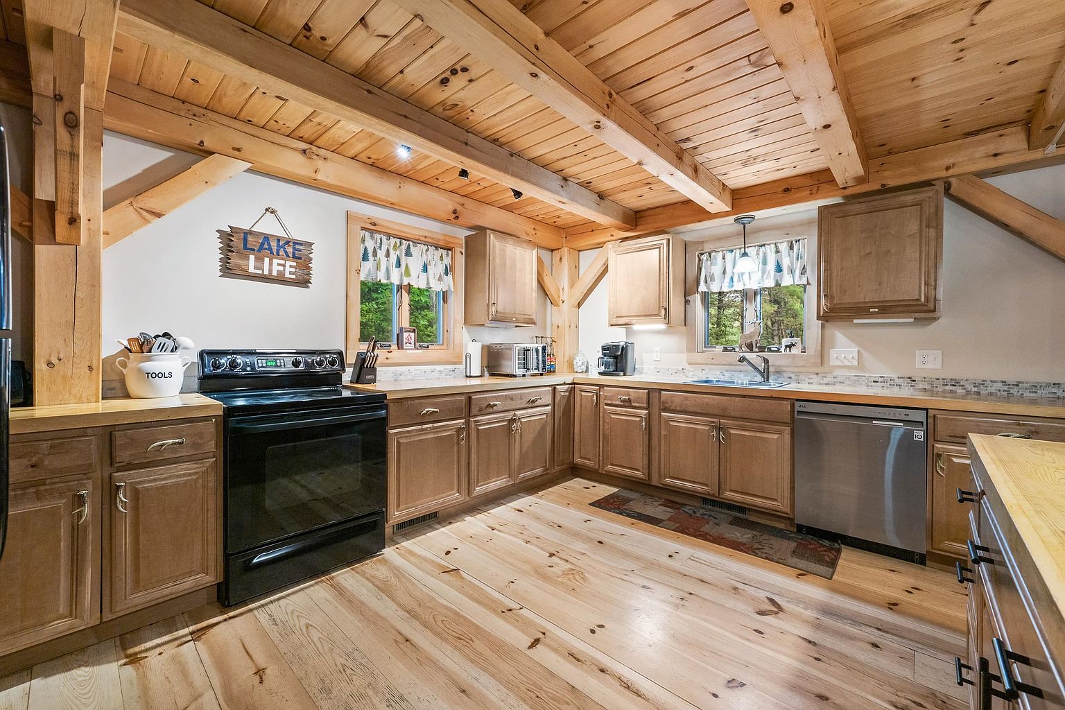 This rustic-style kitchen features warm wood cabinetry, exposed timber ceiling beams, and light-toned wood flooring. The space is equipped with a black electric range, a stainless steel dishwasher, and a neutral-toned backsplash, all illuminated by natural light from two windows adorned with patterned valances. A decorative 'LAKE LIFE' sign adds a charming, thematic touch to the cozy, cabin-inspired atmosphere.