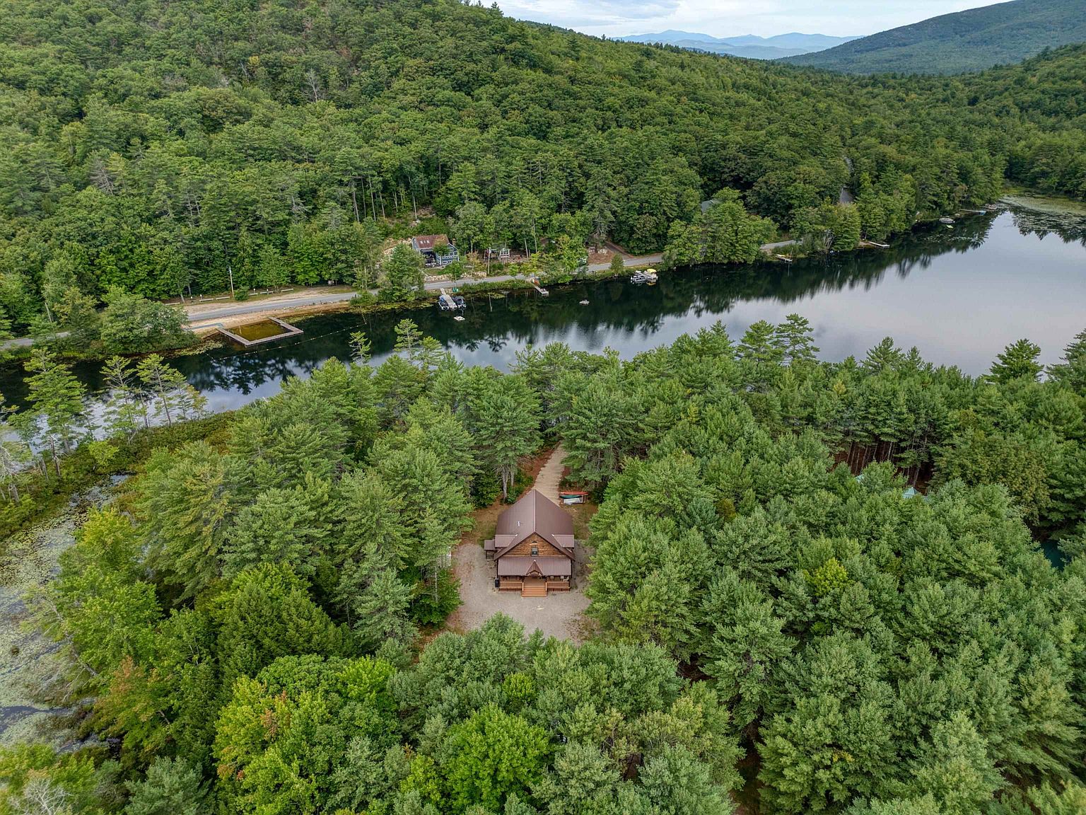 This high-angle aerial shot captures a secluded log cabin nestled within a dense, lush forest. The property features a gravel driveway leading to the home, which is situated near a calm, reflective lake bordered by trees and a quiet road. The perspective emphasizes the serene, private setting and the natural beauty of the surrounding landscape.
