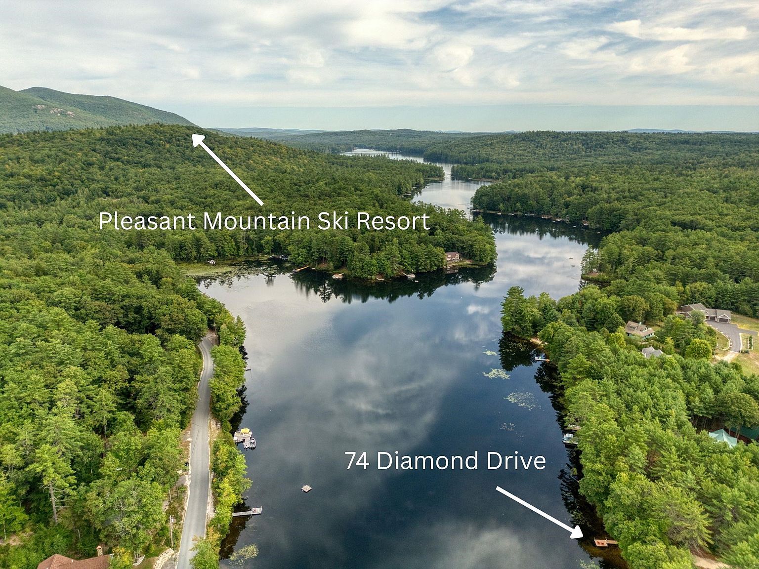 This high-angle aerial perspective captures a serene lakefront setting surrounded by dense, lush forest. The image highlights the property's proximity to the water, featuring a private dock and a winding road, while pointing out the nearby Pleasant Mountain Ski Resort in the distance. The composition emphasizes the tranquil, natural landscape and the secluded nature of the location.