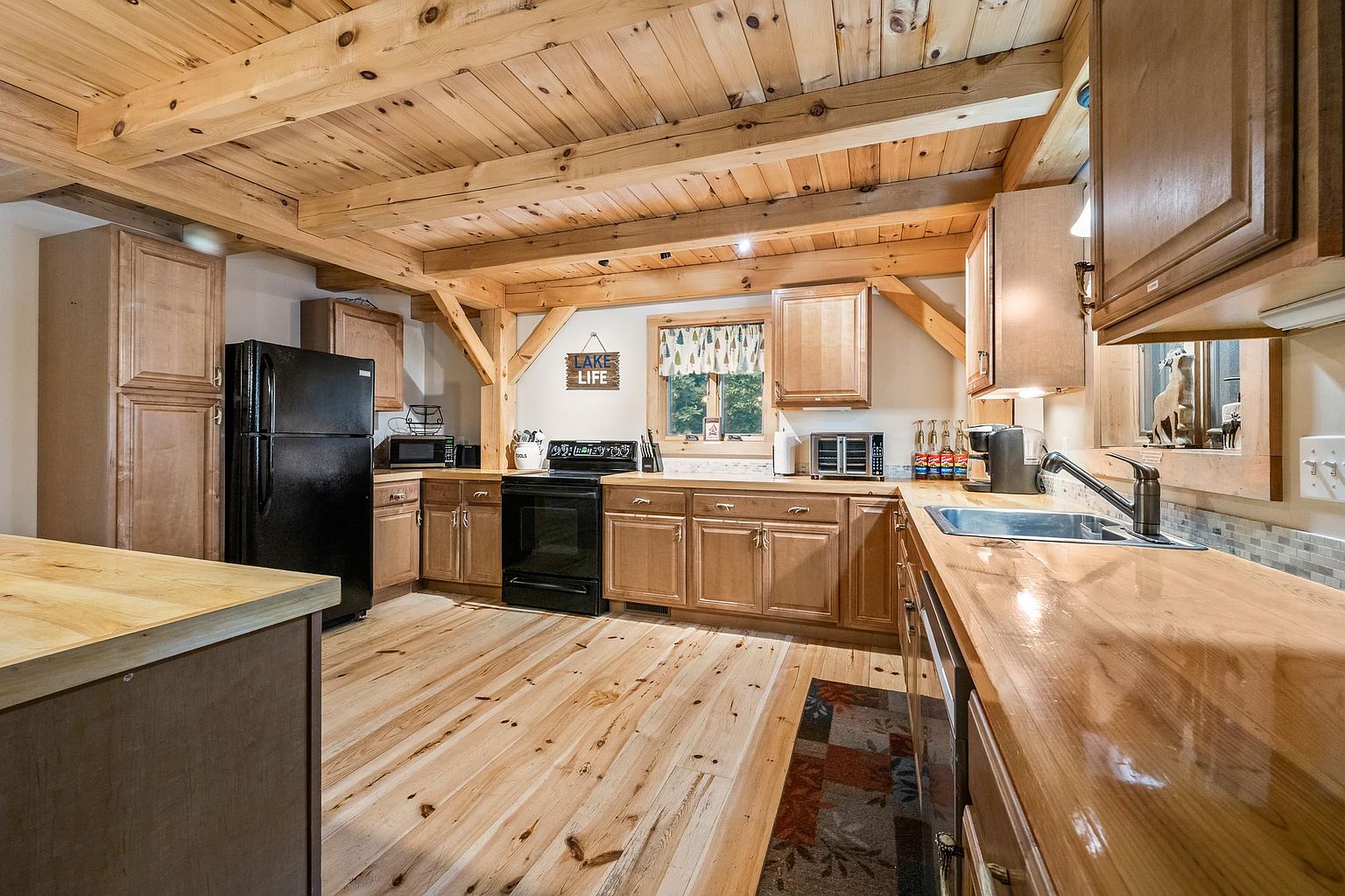 This rustic-style kitchen features warm light wood cabinetry, exposed timber ceiling beams, and matching wood flooring. The space is equipped with black appliances, a polished wood countertop, and a double-basin sink, all set against a neutral wall backdrop. A small window above the counter brings in natural light, while a decorative sign adds a cozy, cabin-like charm to the room.