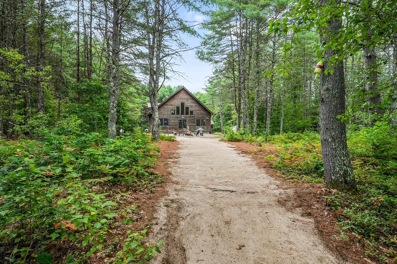 A rustic log cabin is nestled within a dense, lush forest, approached by a wide sandy path that leads directly to the home's entrance. The structure features a prominent gabled roof, a large deck area with outdoor seating, and numerous windows that reflect the surrounding greenery. The scene captures a serene, secluded woodland retreat with a natural, earthy aesthetic.