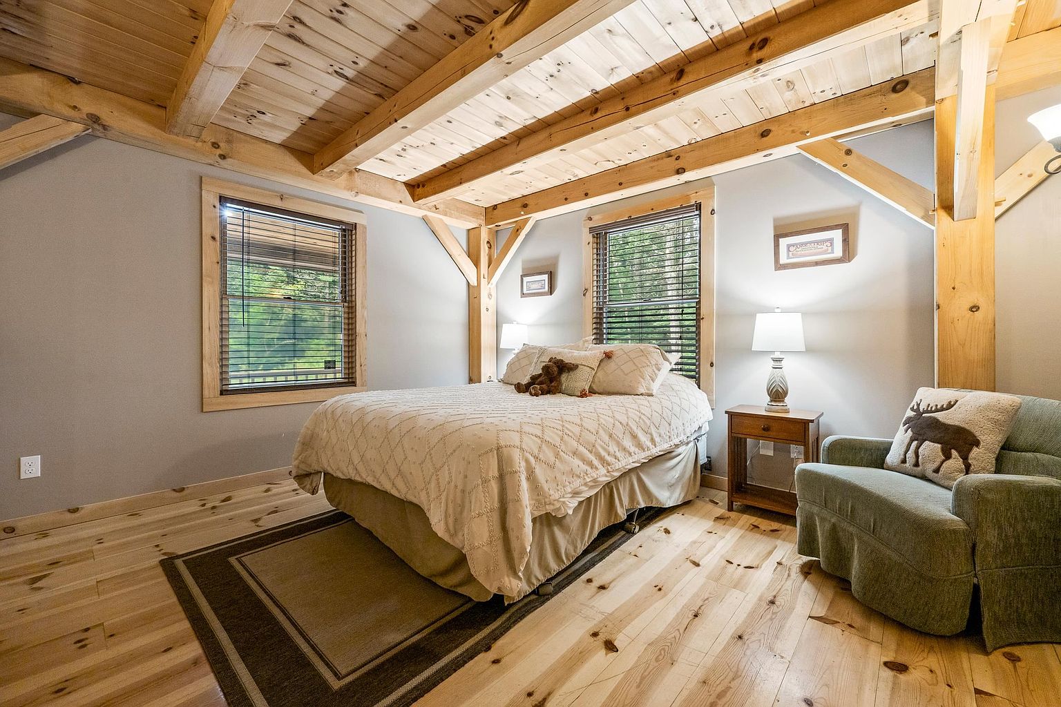 This cozy bedroom features rustic timber-frame construction with exposed wooden beams on the ceiling and walls. The room is furnished with a queen-sized bed, a wooden nightstand with a lamp, and a comfortable armchair adorned with a decorative moose-themed pillow. Large windows with wooden blinds allow natural light to filter in, highlighting the light-toned wood flooring and the warm, cabin-inspired aesthetic.