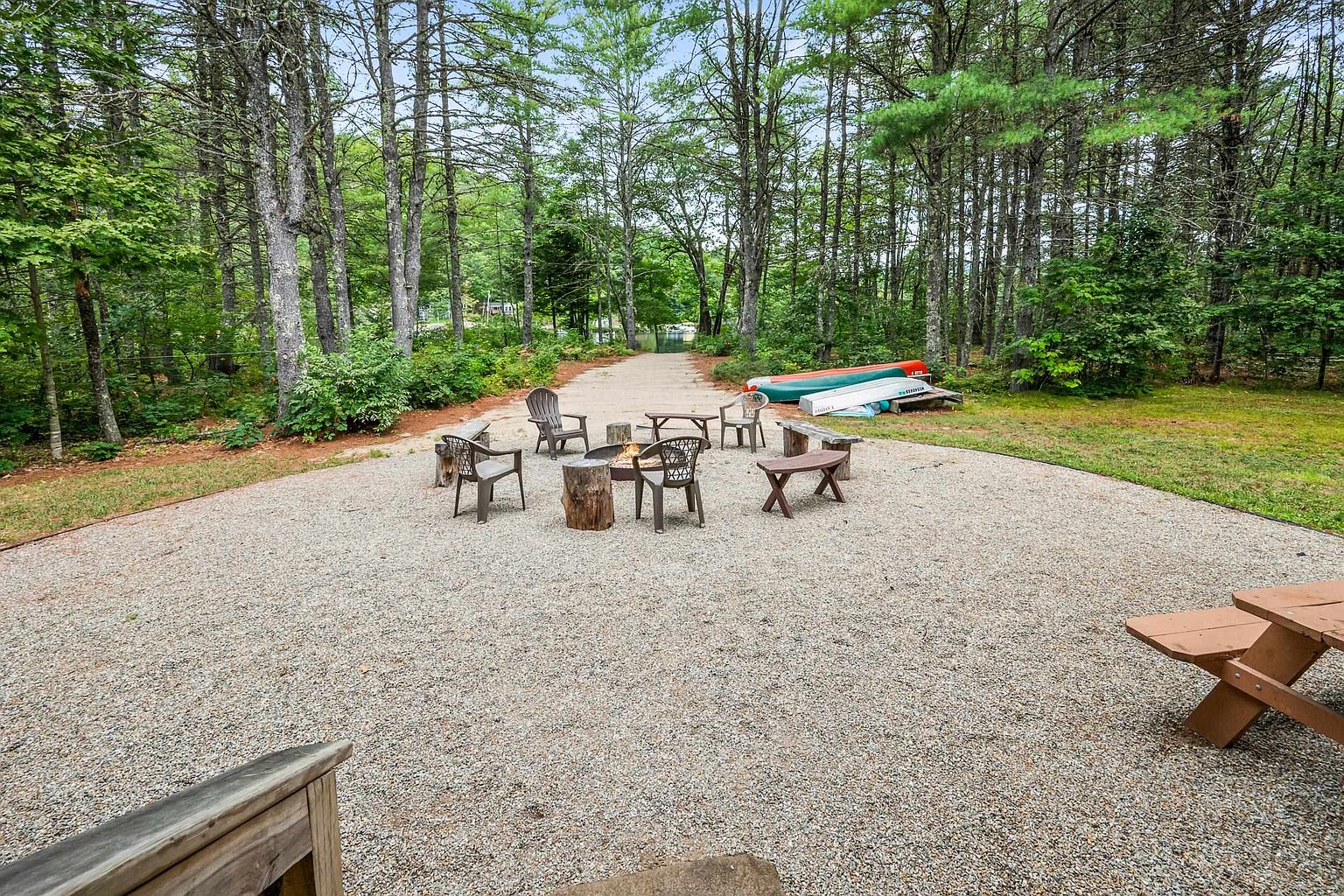This outdoor space features a large, gravel-covered fire pit area furnished with various chairs and a wooden bench, perfect for social gatherings. The area is surrounded by a lush, wooded landscape with a path leading toward a glimpse of a lake in the distance. Two canoes rest on the grass near the trees, enhancing the serene, recreational atmosphere of this property.