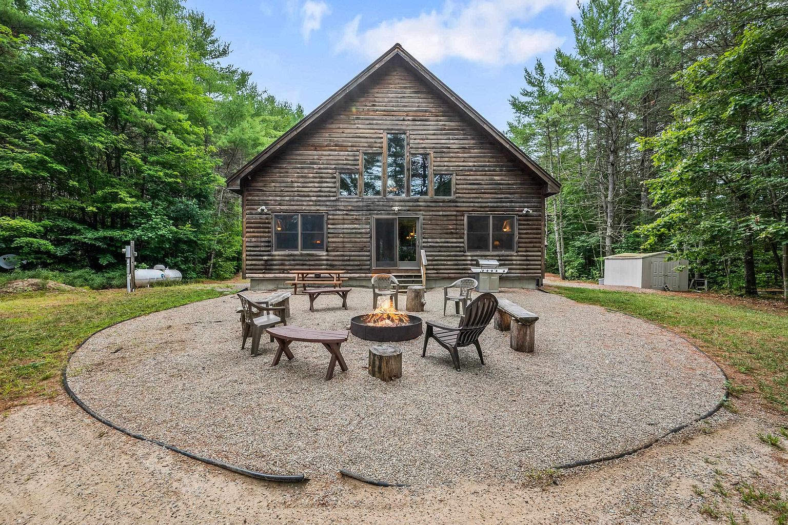 This rear view of a rustic log cabin showcases a spacious gravel fire pit area centered in a wooded backyard. The home features a prominent A-frame design with large windows, a central sliding glass door, and a natural wood exterior that blends into the surrounding forest. The scene is set for outdoor relaxation, with various chairs and a picnic table arranged around a glowing fire pit.