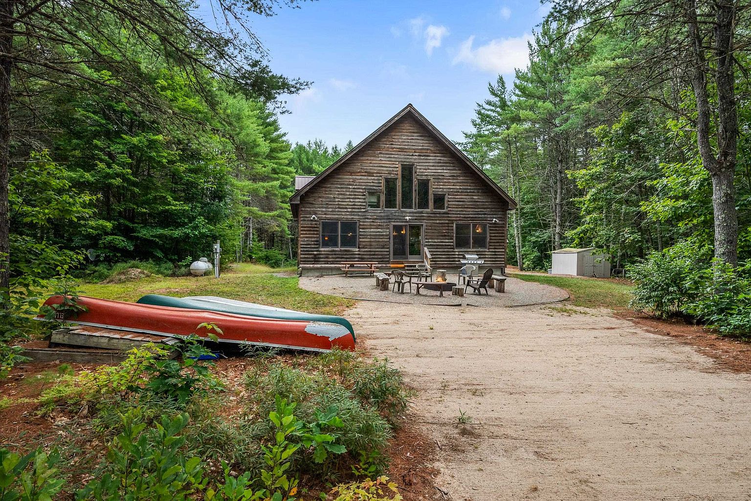 This rustic log cabin is nestled in a lush, wooded setting, featuring a prominent A-frame design with a gravel patio area for outdoor seating. The exterior showcases natural log construction, a central set of glass doors, and a series of windows that allow for natural light. In the foreground, two canoes rest on the sandy ground, emphasizing the property's recreational appeal and serene, secluded atmosphere.