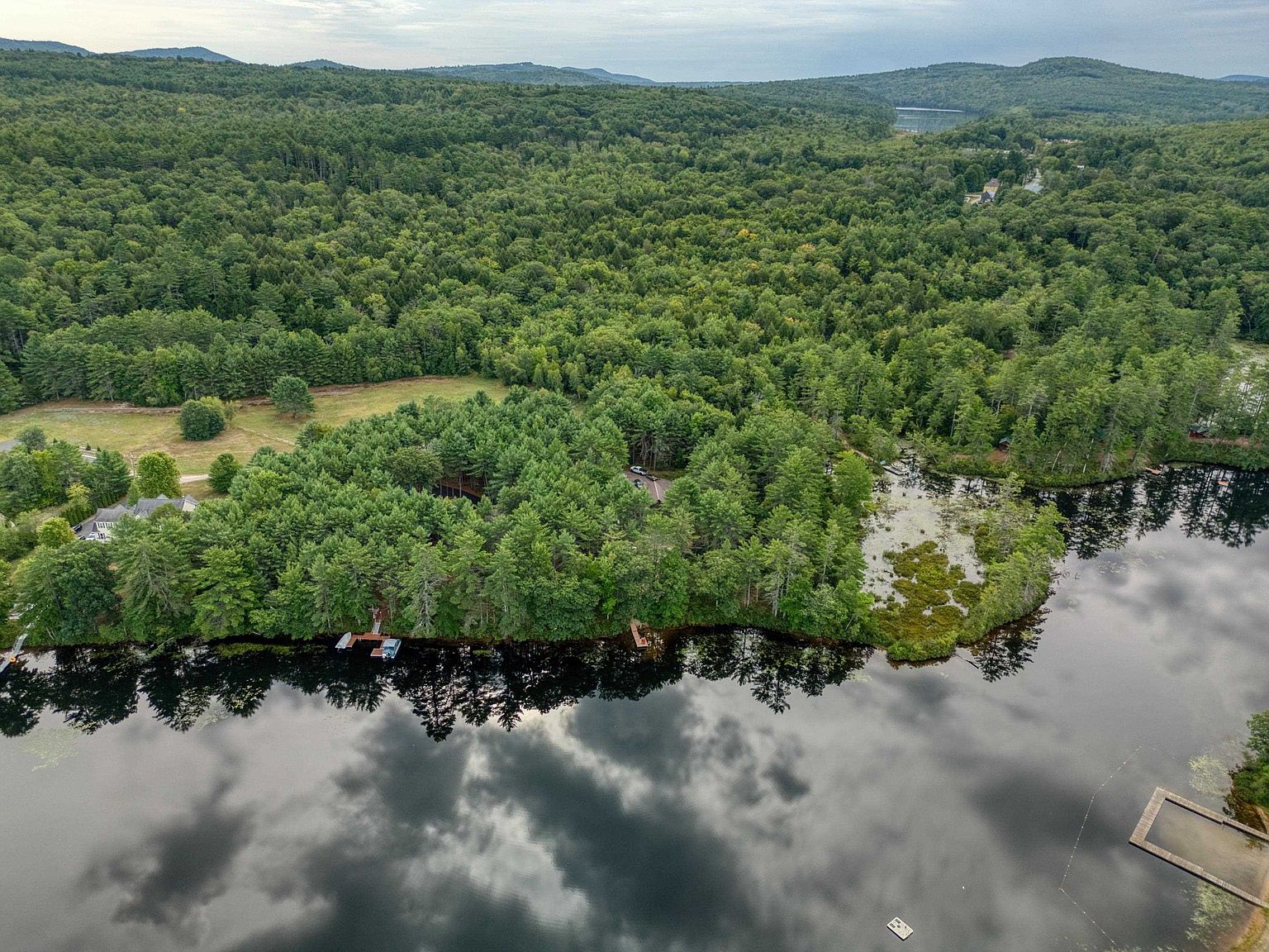 This high-angle aerial perspective captures a serene waterfront property nestled within a dense, lush forest. The scene features a calm lake reflecting the cloudy sky, a private wooden dock extending into the water, and a secluded home partially obscured by mature trees. The expansive landscape highlights the property's peaceful, natural setting and direct access to the water.