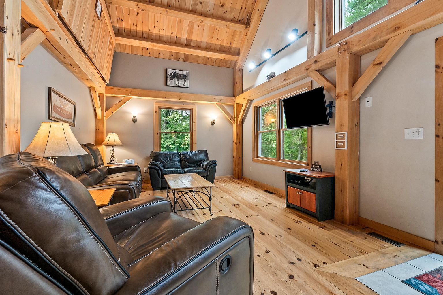 This rustic living room features a vaulted ceiling with exposed timber beams and light wood flooring, creating a warm and inviting cabin atmosphere. The space is furnished with dark leather seating, including a recliner in the foreground and a sofa against the back wall, centered around a simple coffee table. Large windows allow natural light to fill the room, which is accented by wall-mounted lighting and a television mounted above a small media console.