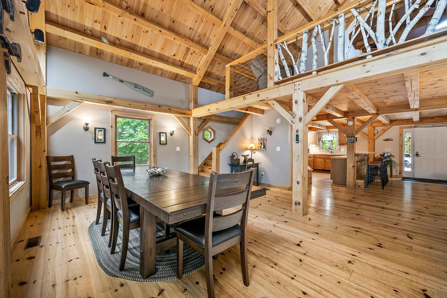 This rustic, open-concept dining area features a large wooden table surrounded by matching chairs, set upon a textured oval rug. The space is defined by exposed timber beams, light wood flooring, and a lofted area above with a unique birch branch railing. A view into the adjacent kitchen and a bright entryway adds to the airy, cabin-inspired atmosphere of this home.