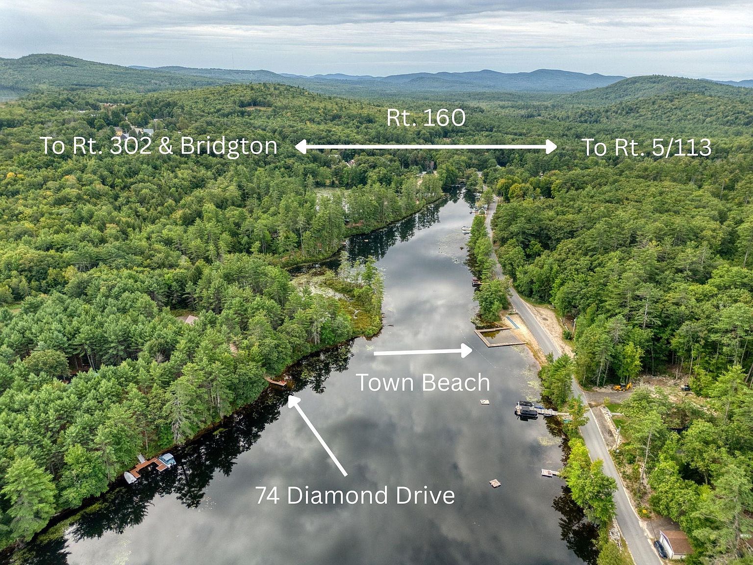 This high-angle aerial view captures a serene lakefront setting, showcasing the natural landscape and proximity to local landmarks. The image highlights the waterfront property at 74 Diamond Drive, the nearby town beach, and the surrounding dense forest canopy. The perspective provides a clear sense of the property's location relative to the main road, Rt. 160, and the broader regional geography.