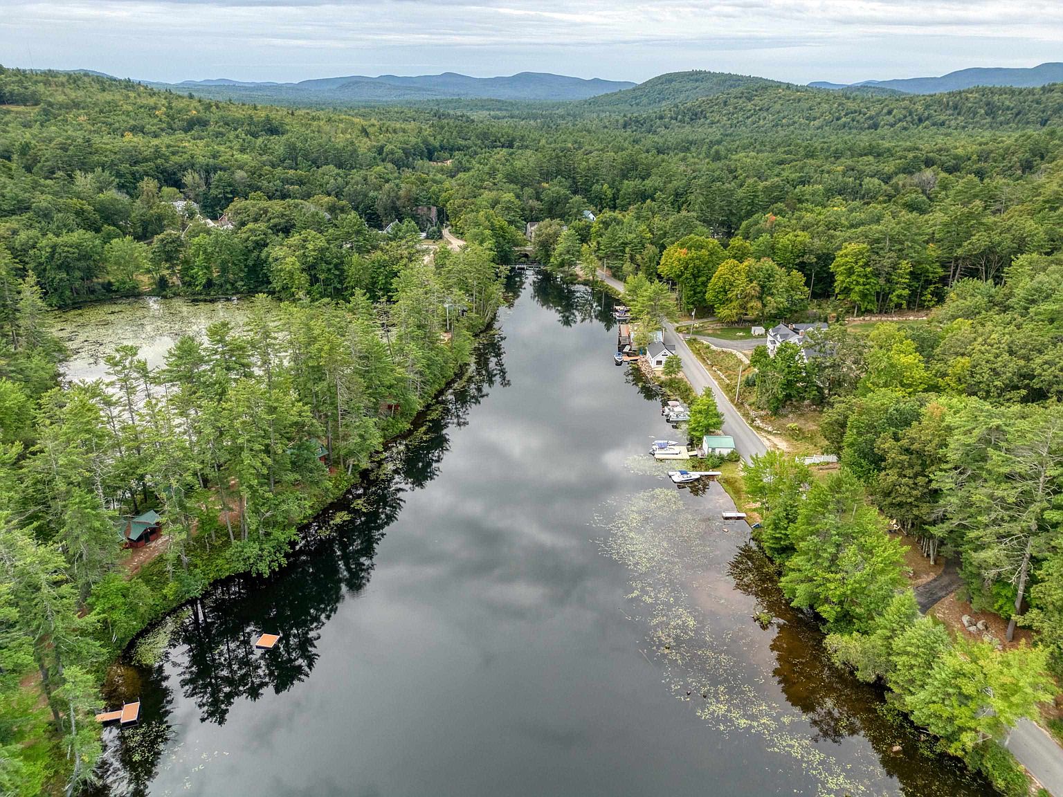 This high-angle aerial perspective captures a serene lakefront setting surrounded by dense, lush forest and rolling hills in the distance. The scene features a calm body of water reflecting the cloudy sky, with a quiet road running along the shoreline dotted with small cottages, private docks, and moored boats. The composition emphasizes the peaceful, natural environment and the recreational potential of the waterfront property.