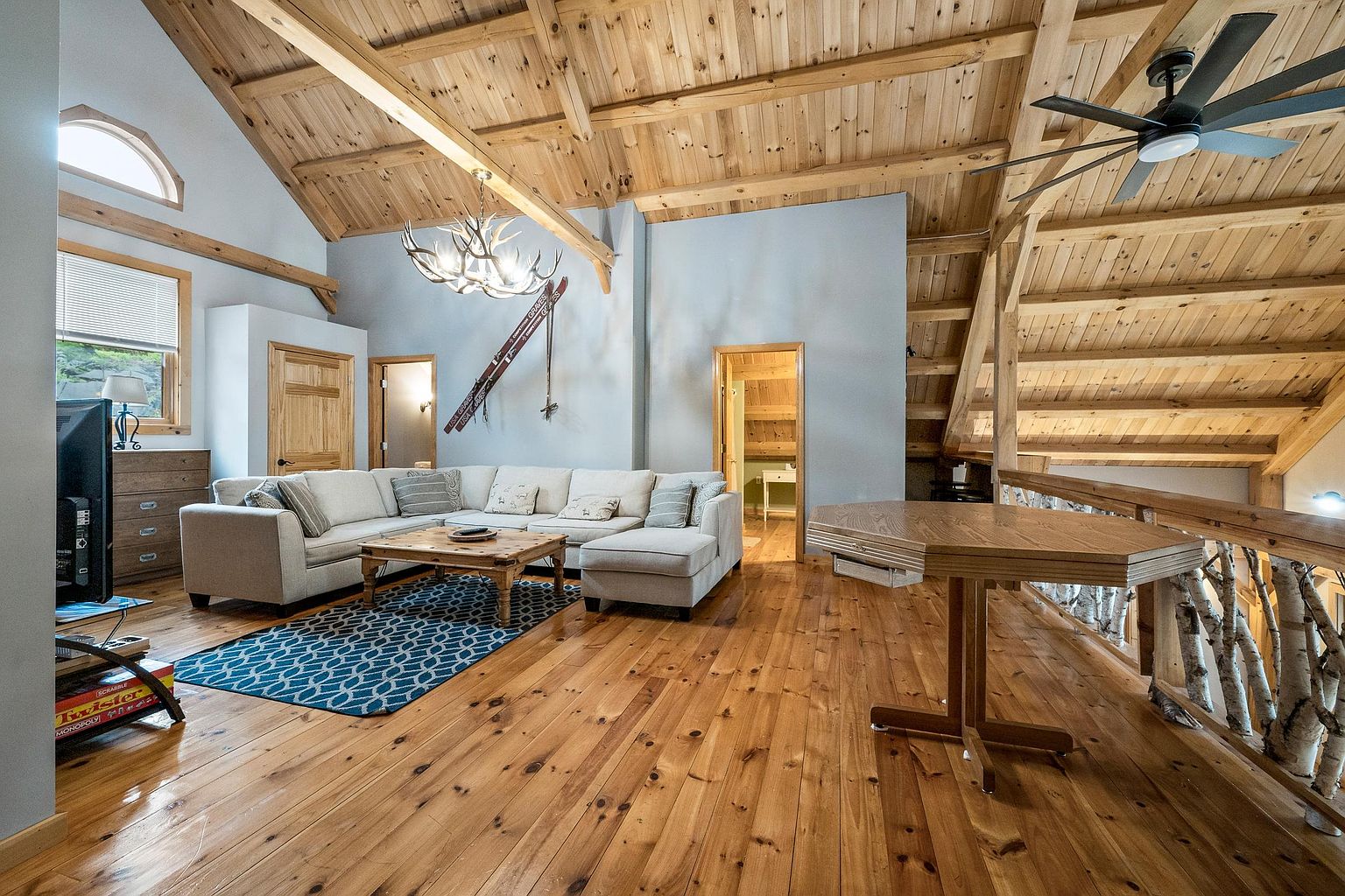 This spacious, open-concept living area features a vaulted ceiling with exposed wooden beams and a rustic, lodge-inspired aesthetic. A large, light-colored sectional sofa sits atop a patterned area rug, centered around a wooden coffee table, while a unique antler chandelier hangs above. The room includes a wooden table in the foreground, a ceiling fan, and a view into an adjacent hallway, all set against a backdrop of warm wood flooring and light-toned walls.