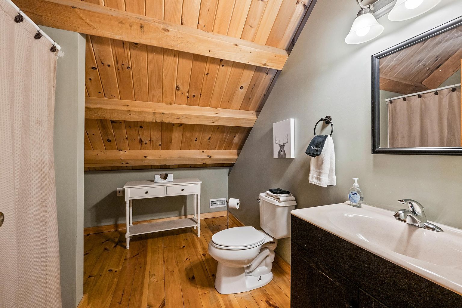 This rustic-style bathroom features a slanted wood-paneled ceiling with exposed beams, creating a cozy, cabin-like atmosphere. The space includes a dark vanity with a light-toned sink, a white toilet, and a small console table tucked under the eaves. A framed mirror hangs above the vanity, and a small piece of deer-themed wall art adds a decorative touch to the neutral-toned walls.