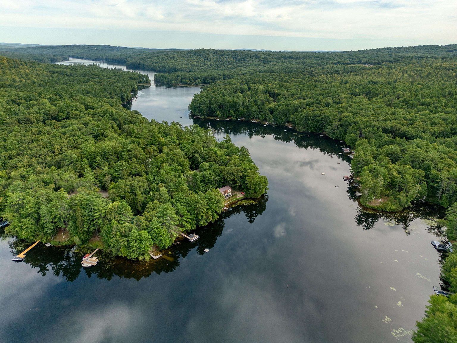 This high-angle aerial perspective captures a serene lakefront property nestled within a dense, lush forest. The scene highlights the natural beauty of the landscape, featuring a calm body of water reflecting the sky, several private docks, and a secluded cabin tucked among the trees. The composition emphasizes the peaceful, private nature of the setting, making it an ideal retreat for those seeking waterfront living.