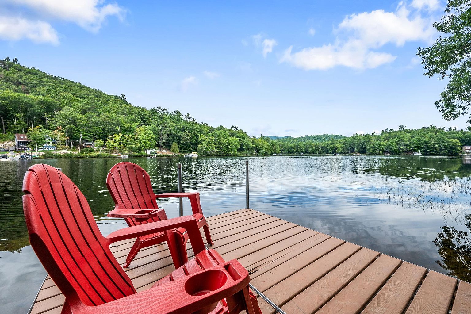 This image captures a serene lakeside view from a wooden dock, featuring two bright red Adirondack chairs positioned to overlook the calm water. The scene is framed by a lush, tree-covered hillside and a clear blue sky with scattered clouds, creating a peaceful and inviting atmosphere. The perspective is from the dock looking out toward the center of the lake, emphasizing the tranquil waterfront lifestyle.