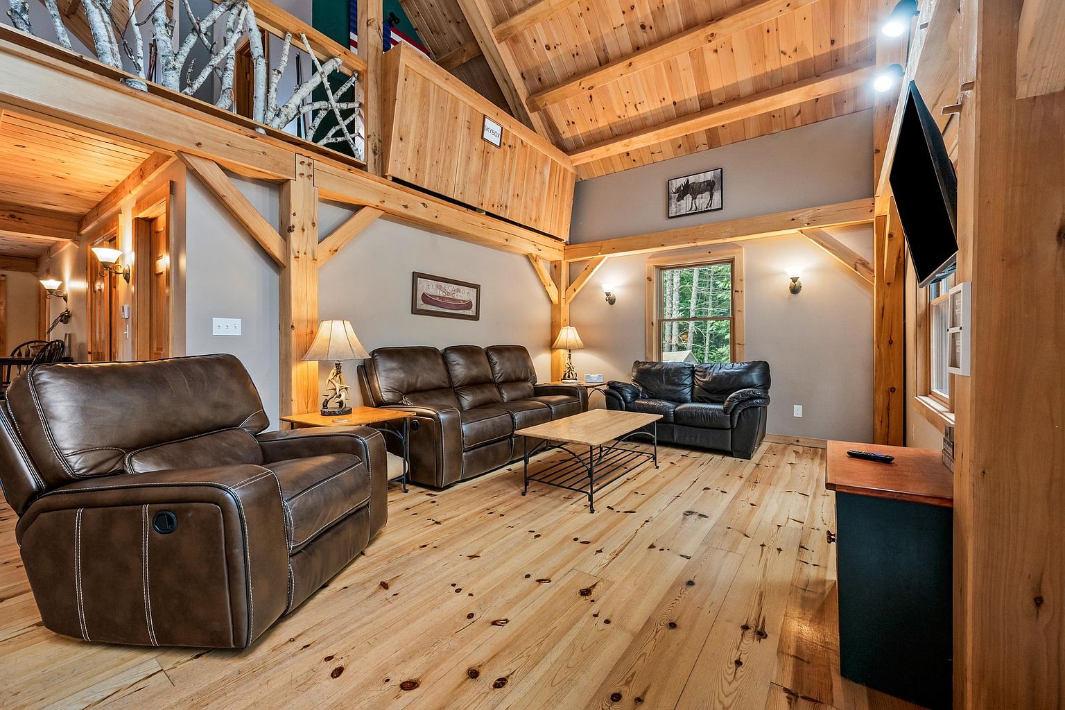 This rustic living room features warm wood-paneled walls and ceilings with exposed timber beams, creating a cozy cabin atmosphere. The space is furnished with a leather recliner, a matching sofa, and a smaller loveseat arranged around a central coffee table, all set upon light-toned wood flooring. A loft area with a unique birch-branch railing overlooks the room, while a window provides a view of the surrounding trees, enhancing the natural, lodge-like aesthetic.