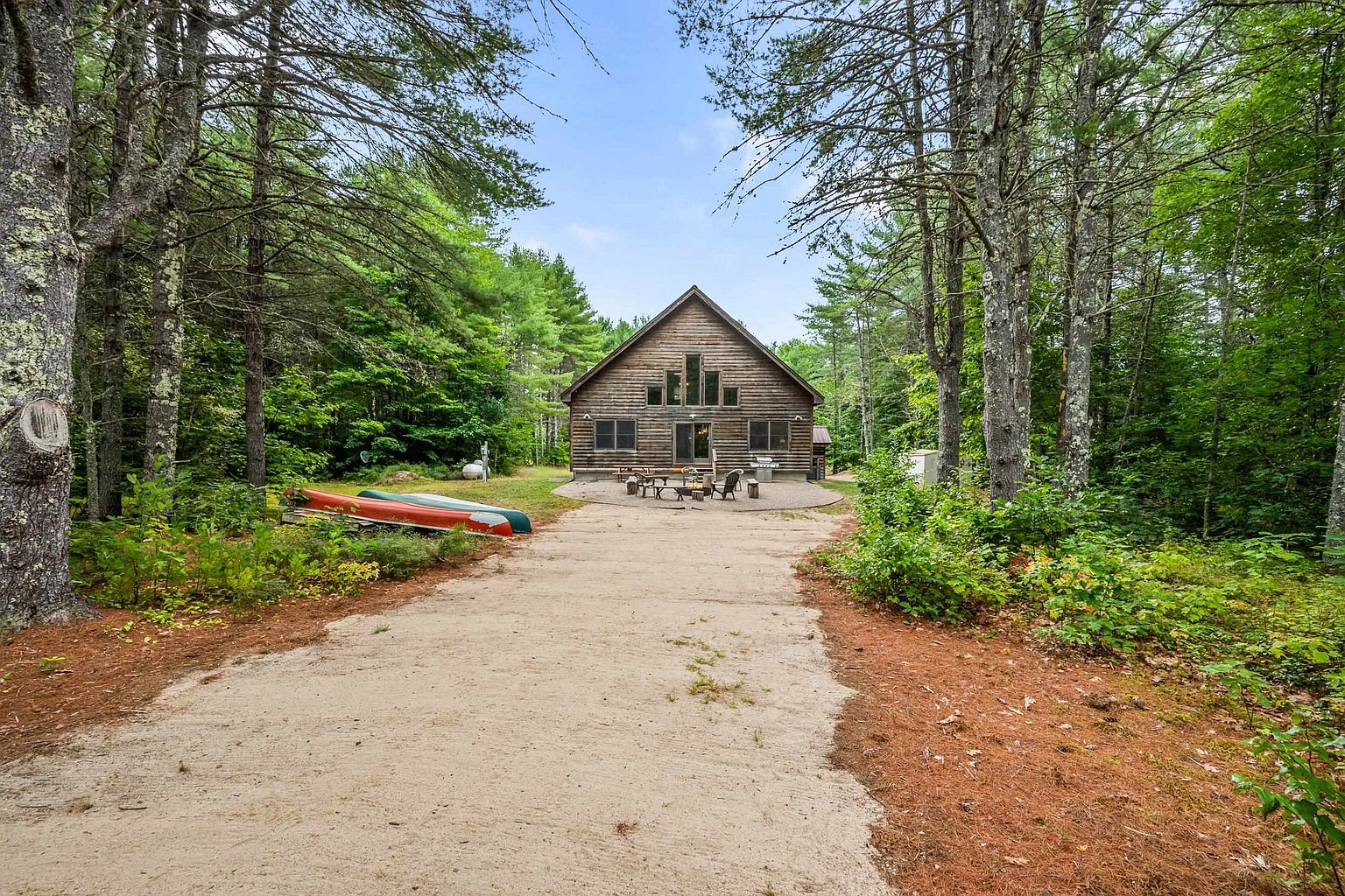 This image captures the rear exterior of a rustic log cabin nestled within a dense, wooded forest. A sandy path leads toward a circular stone patio area equipped with outdoor seating and a fire pit, while two canoes rest on the grass to the left. The cabin features a prominent A-frame design with large windows that reflect the surrounding trees, creating a serene and secluded atmosphere.