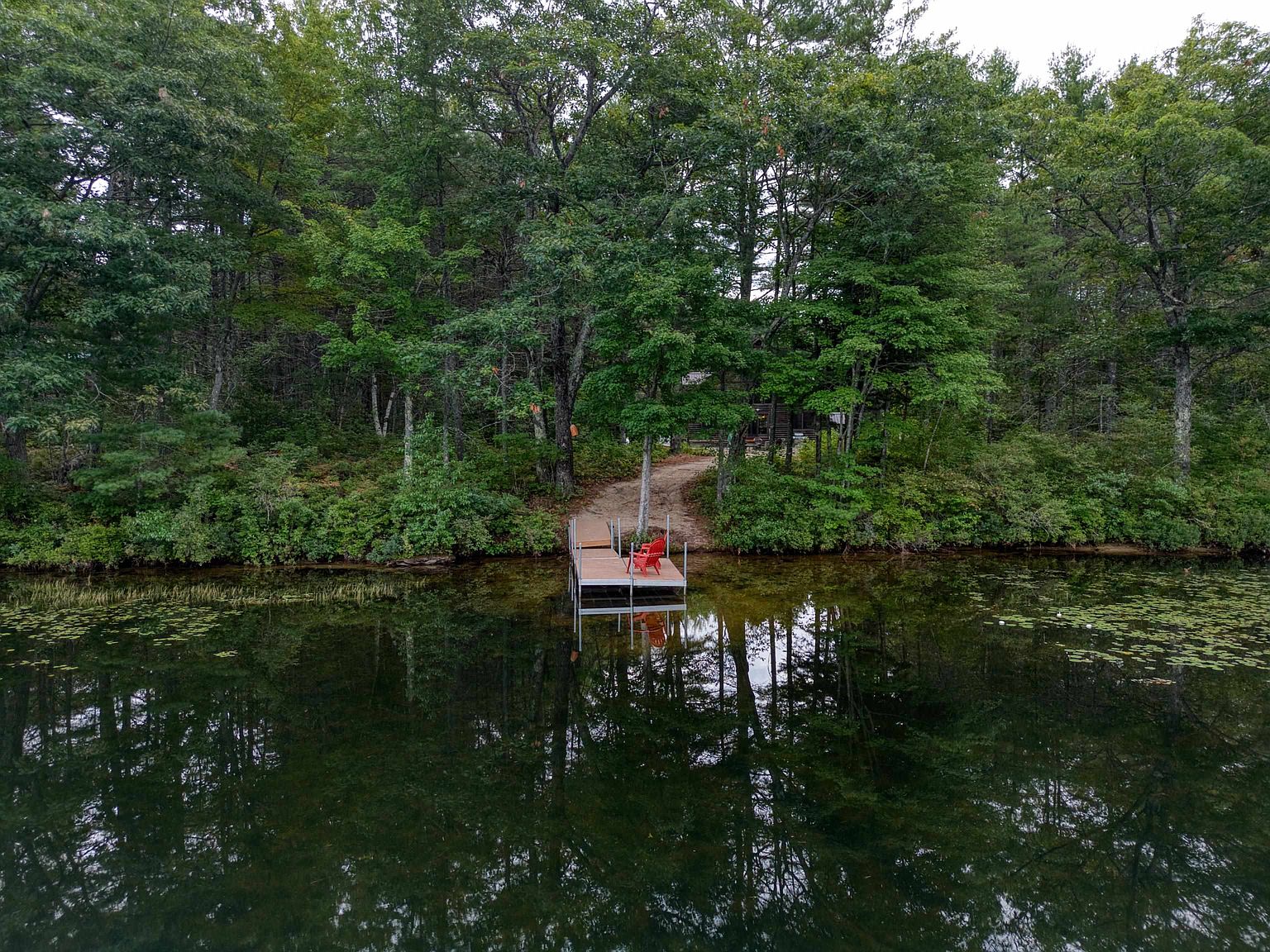 This aerial perspective captures a serene lakeside setting featuring a wooden dock extending into calm, reflective water. A small, bright red chair sits on the dock, providing a vibrant focal point against the lush, dense forest backdrop. A dirt path leads from the water's edge into the trees, suggesting a secluded and peaceful retreat.