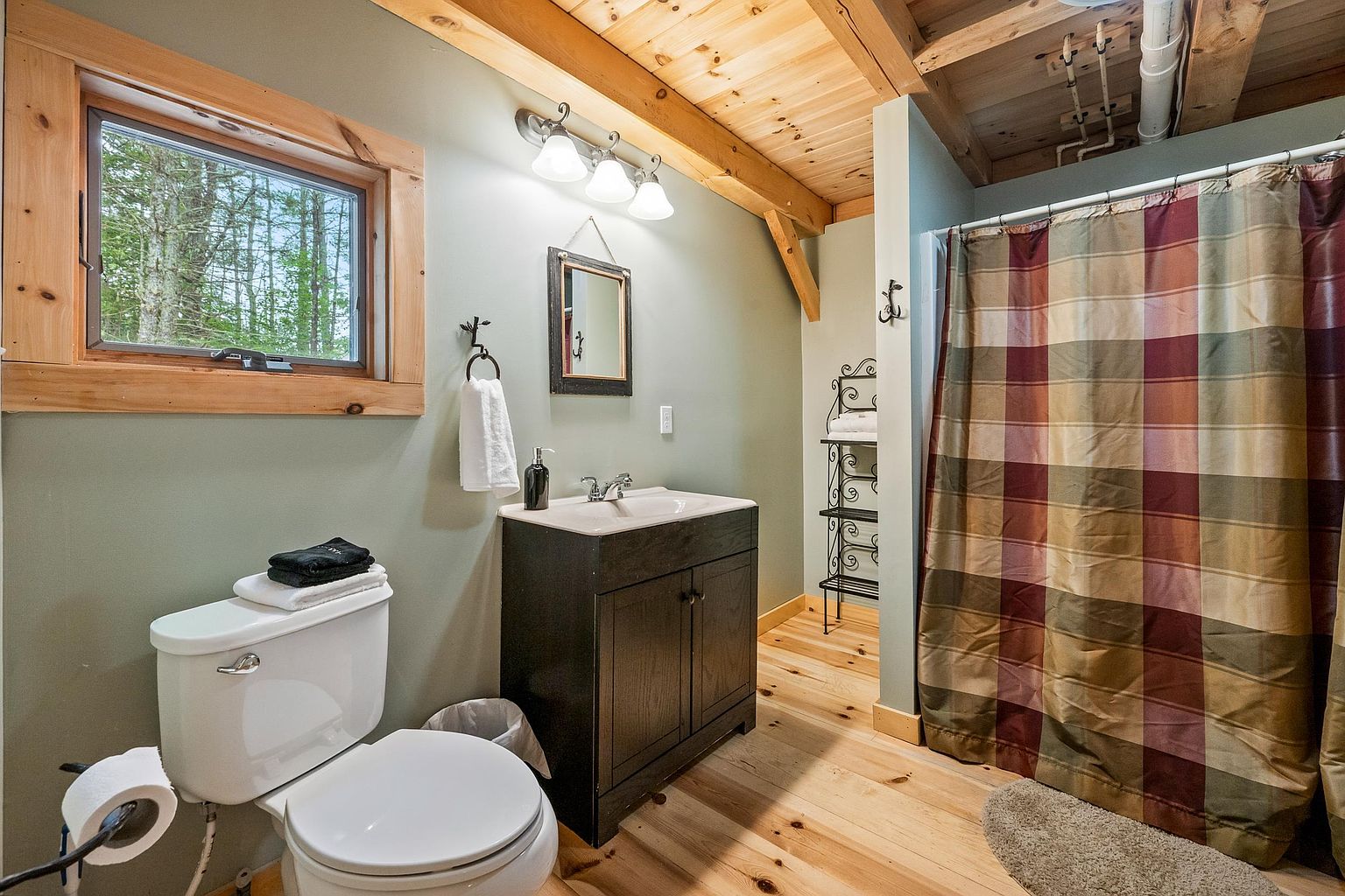 This rustic-style bathroom features light wood flooring and exposed wooden ceiling beams, creating a warm, cabin-like atmosphere. The space includes a dark wood vanity with a light-colored sink, a toilet, and a shower area enclosed by a plaid curtain. A window with a natural wood frame provides a view of the surrounding trees, while a metal shelving unit offers storage for towels.