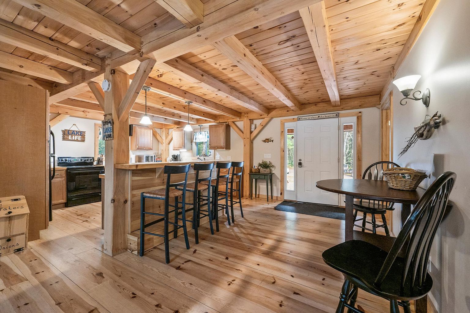 This rustic-style kitchen and dining area features exposed timber beams and light wood flooring, creating a warm, cabin-like atmosphere. The space includes a breakfast bar with four black stools, a functional kitchen area with dark appliances, and a small dining nook near the front entryway. The open-concept layout is illuminated by pendant lighting and natural light from the front door, offering a cozy and inviting living space.