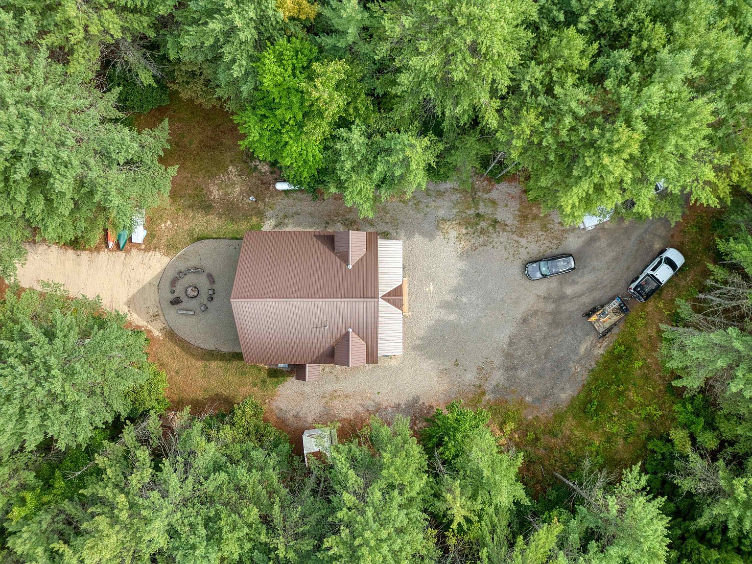 This top-down aerial view captures a secluded property nestled within a dense, lush forest. The home features a prominent brown metal roof and is surrounded by a gravel driveway, a circular stone fire pit area, and ample parking space for multiple vehicles, including a truck with a trailer. The perspective emphasizes the home's private, wooded setting and its integration into the natural landscape.