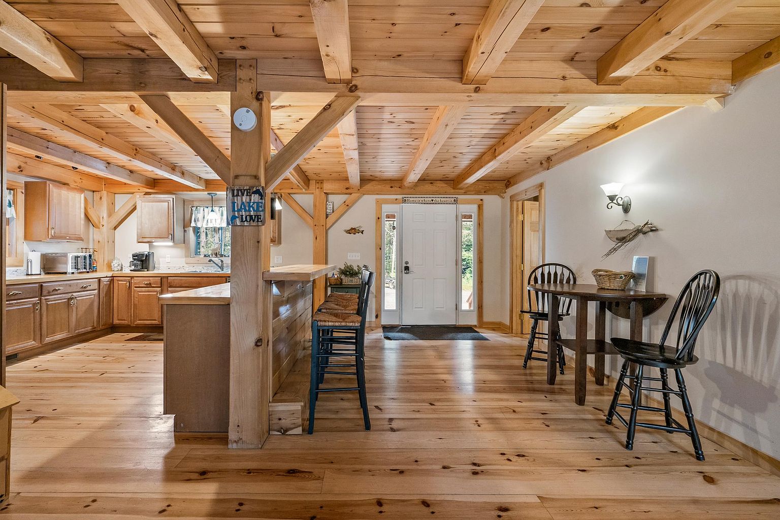 This open-concept kitchen and dining area features rustic timber-frame construction with exposed wooden beams and light-toned wood flooring. The kitchen is equipped with wood cabinetry and a breakfast bar with stools, while a small dining table sits to the right near the front entryway. The perspective is a wide-angle shot from the kitchen looking toward the front door, highlighting the warm, cabin-like aesthetic of the home.