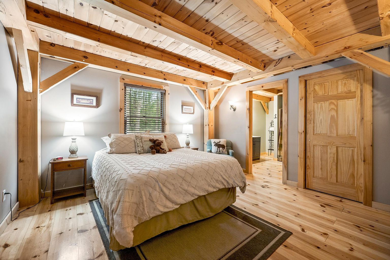 This rustic-style bedroom features exposed timber ceiling beams and light-toned wood flooring, creating a warm and inviting atmosphere. A central bed with a textured white quilt is flanked by two matching nightstands with lamps, while a small armchair sits in the corner near a doorway leading to an adjacent room. The room is accented with natural wood trim around the windows and doors, emphasizing a cozy, cabin-inspired aesthetic.