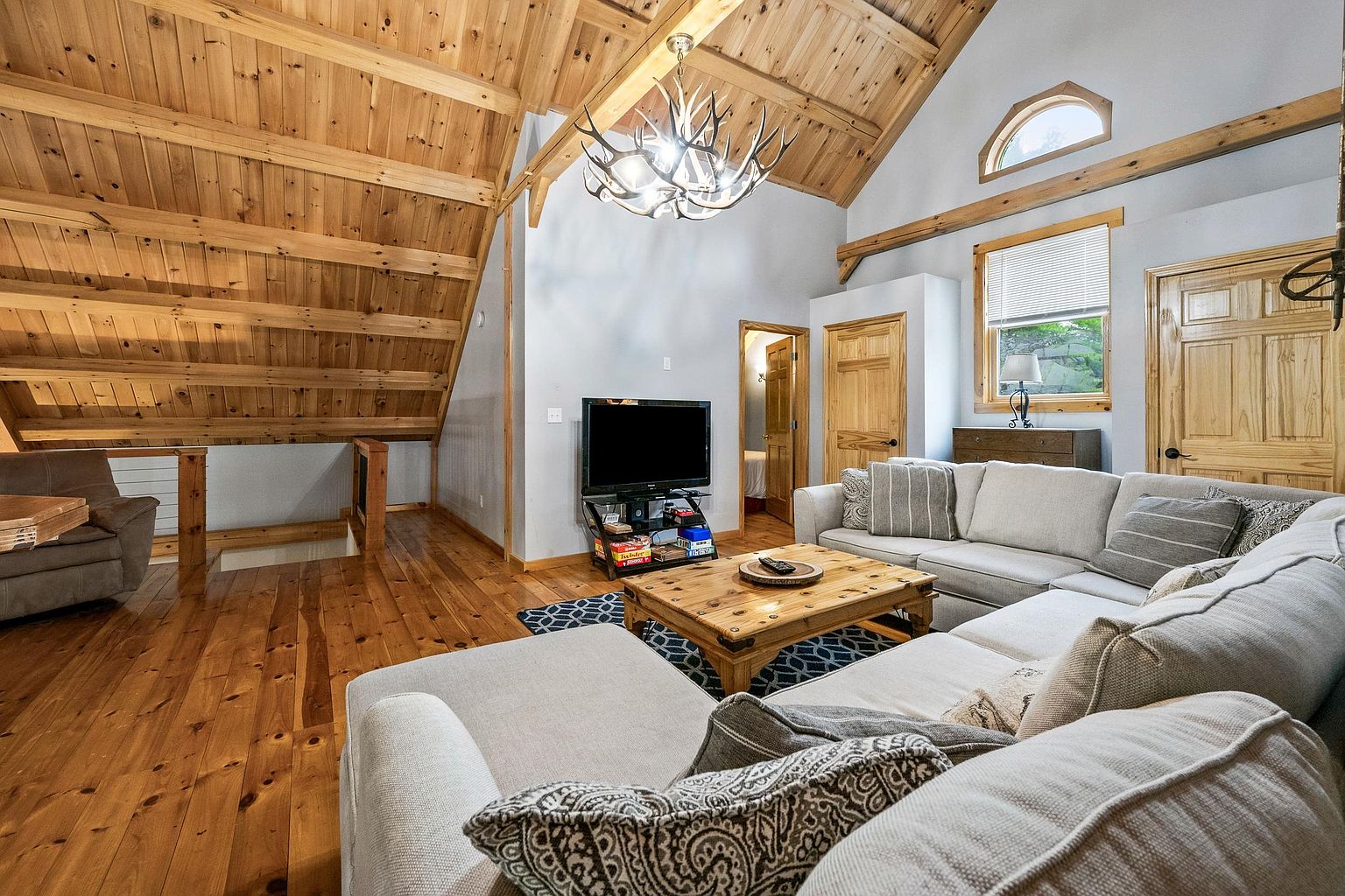 This cozy, rustic-style living room features a vaulted ceiling with exposed wooden beams and a distinctive antler chandelier. The space is furnished with a large, light-colored sectional sofa and a wooden coffee table, all set upon a patterned area rug. A television sits on a stand in the center, while the room's open layout overlooks a staircase leading to the lower level, creating an inviting and airy atmosphere.