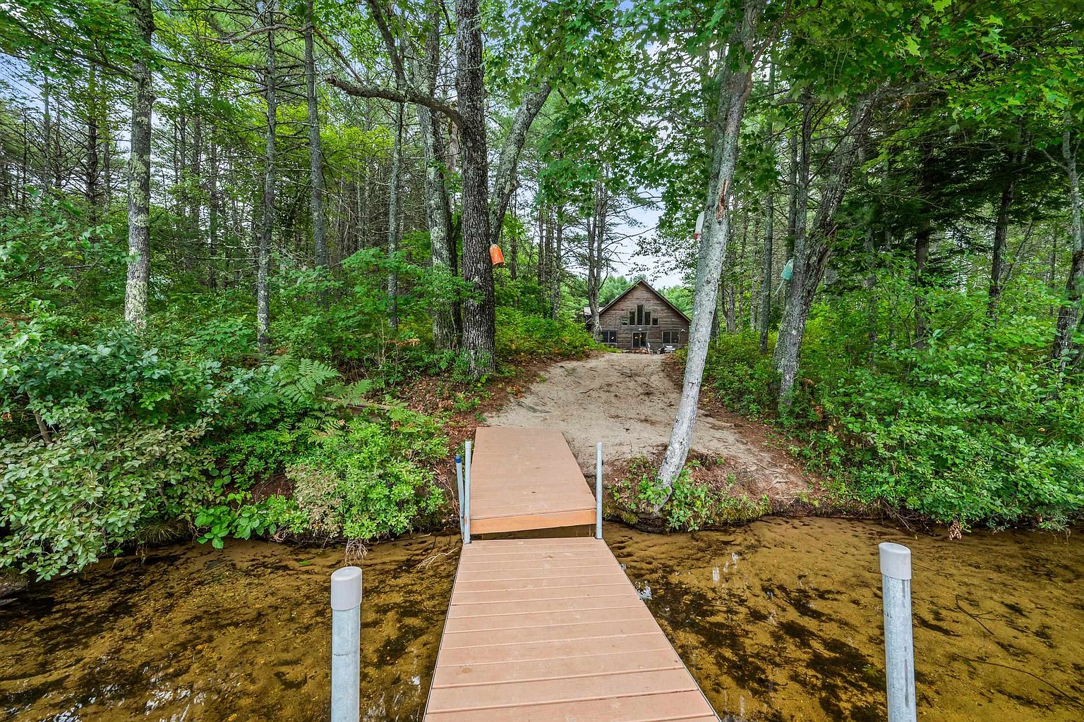 This perspective captures a rustic wooden cabin nestled within a dense, lush forest, viewed from the vantage point of a floating dock on a clear lake. A sandy path leads from the water's edge up toward the home, which features large windows and a natural wood exterior that blends seamlessly into the surrounding trees. The scene evokes a peaceful, secluded retreat atmosphere, highlighting the property's direct waterfront access.