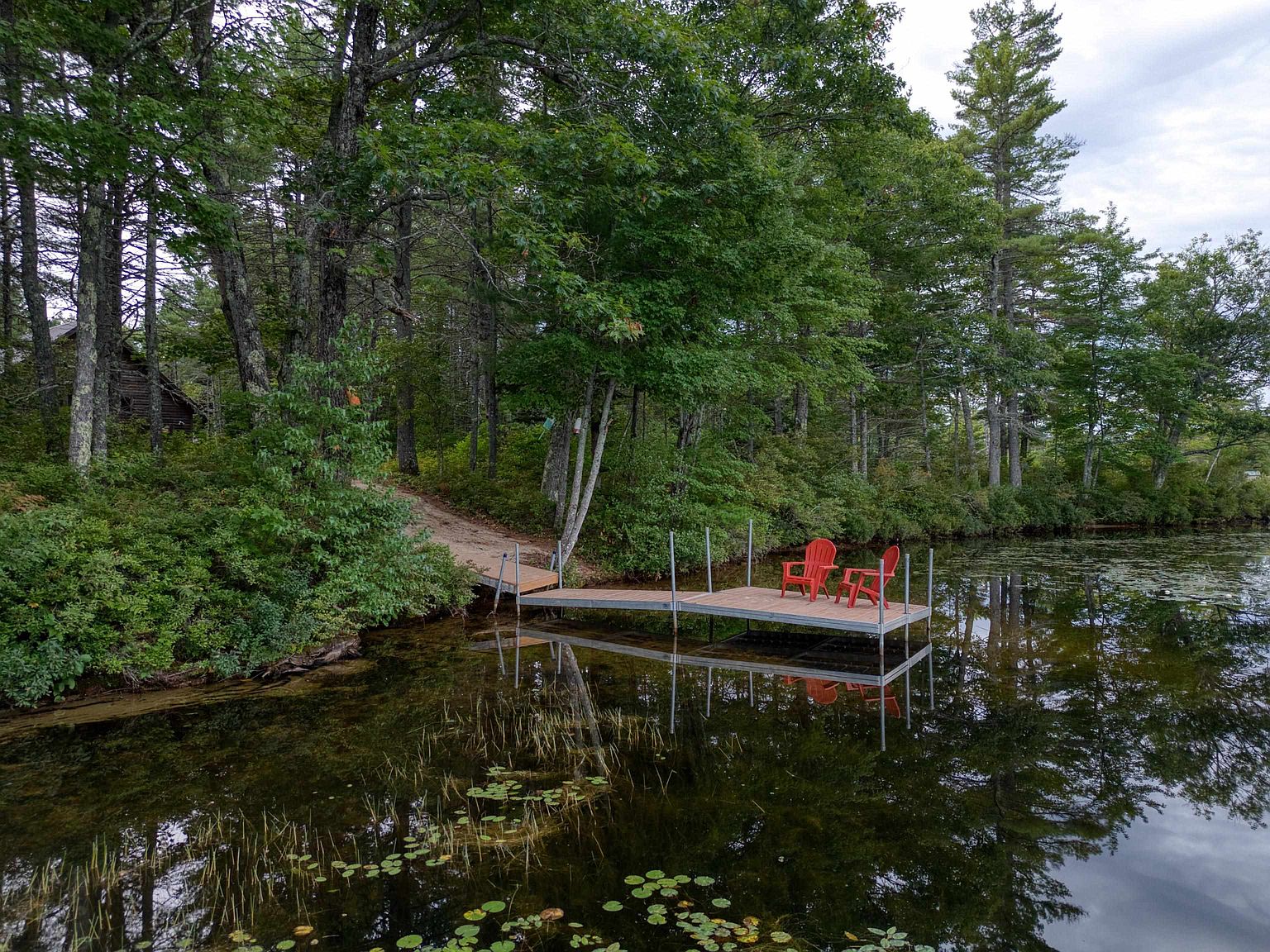 This serene waterfront scene features a floating dock extending from a wooded shoreline into a calm, lily-pad-dotted lake. Two vibrant red Adirondack chairs sit on the dock, providing a peaceful spot to enjoy the natural surroundings. The perspective is captured from a slightly elevated angle, emphasizing the lush forest backdrop and the tranquil, reflective water.