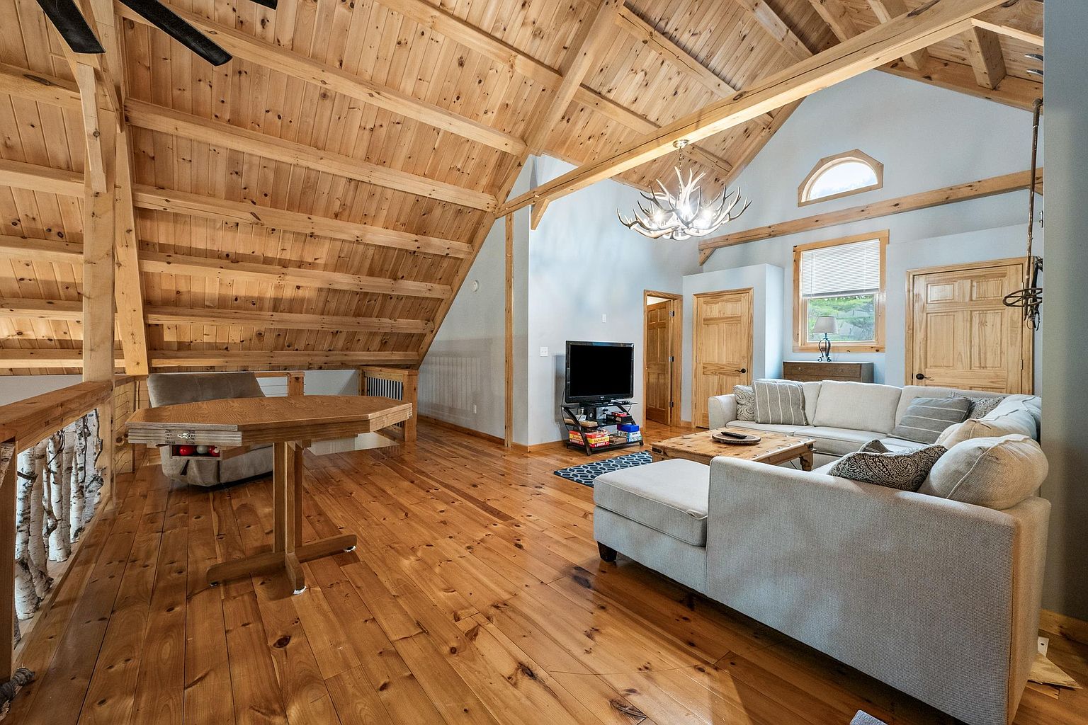 This spacious loft-style living area features a vaulted ceiling with exposed wooden beams and warm wood paneling throughout. The room is furnished with a large, light-colored sectional sofa and a wooden coffee table, creating a cozy gathering space. A unique antler chandelier serves as a focal point, while a game table sits near the open railing overlooking the lower level.