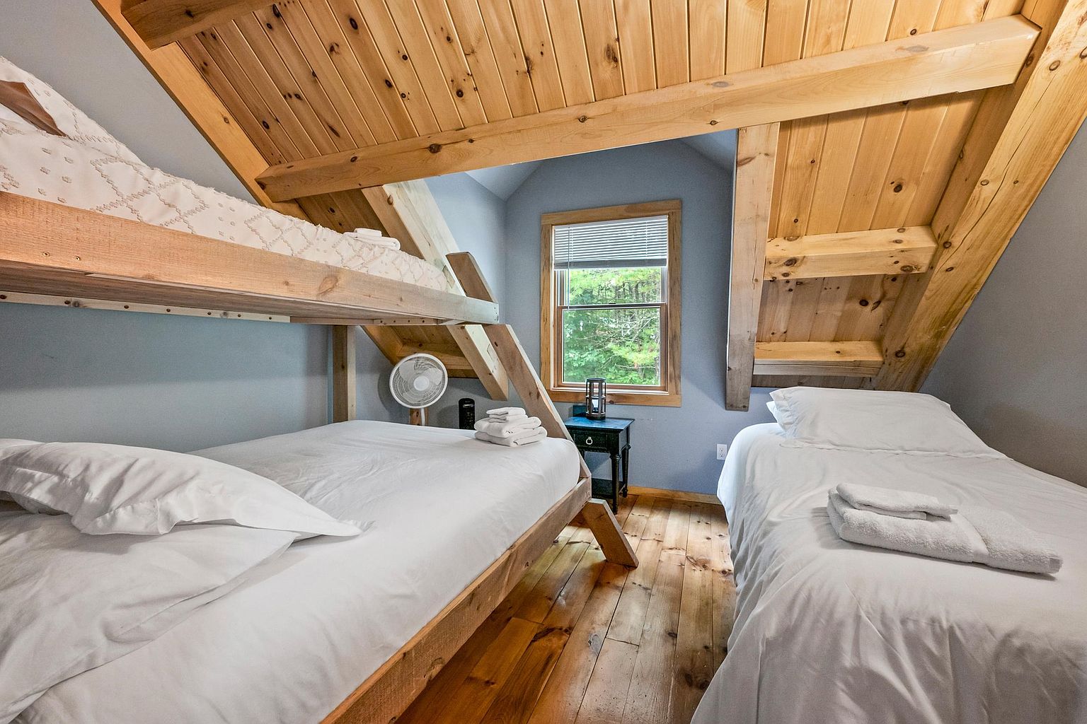 This cozy attic bedroom features a rustic aesthetic with exposed light wood beams and a vaulted ceiling. The room is furnished with a bunk bed on the left and a separate twin bed on the right, both dressed in crisp white linens. A small window centered on the back wall provides natural light, while a dark side table and a portable fan complete the functional, cabin-style space.